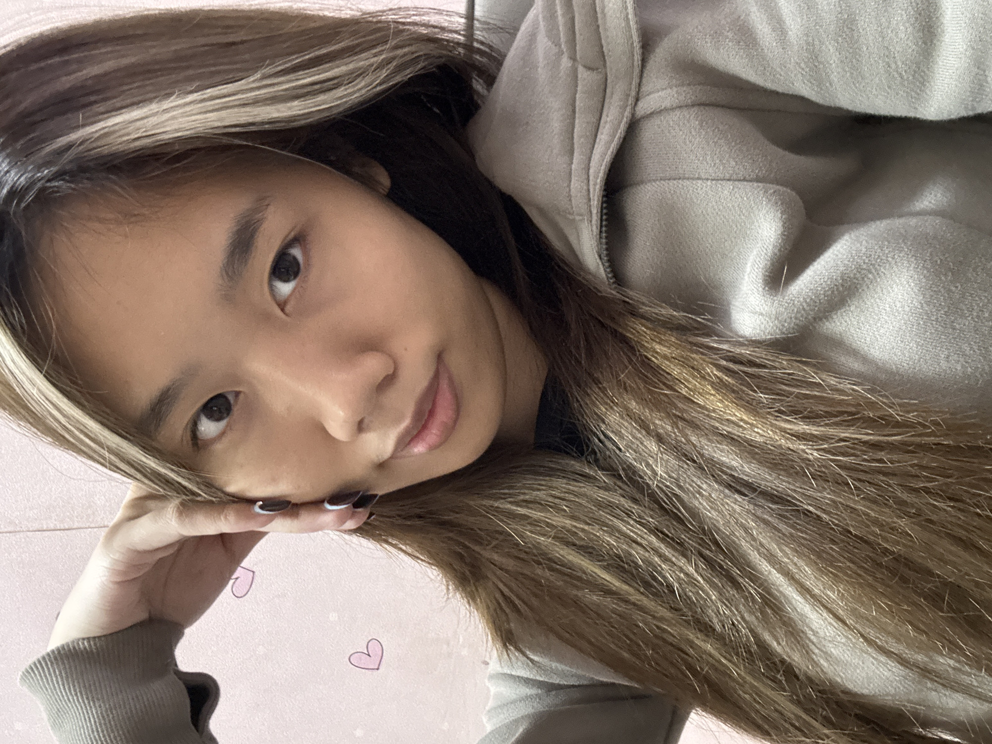 A young woman with long, highlighted brown hair is posing indoors. She is resting her head on her hand. She is wearing a hooded sweatshirt. There are small, cartoonish hearts on the wall behind her. The location in Shapingba District, China is not discernible from the image.