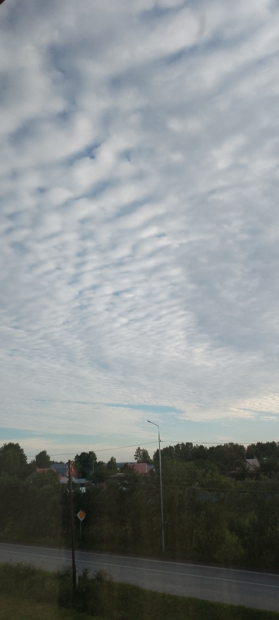 The image depicts a rural scene in Абалак, Russia, under a sky filled with a unique pattern of altocumulus clouds, often described as a "mackerel sky." The clouds are white and fluffy, forming distinct, rippling bands across the pale blue sky. This cloud formation suggests fair weather with some atmospheric instability.
Below the sky, a dense line of dark green trees stretches across the middle ground, obscuring much of the landscape behind them. Peeking through the trees are the roofs of several houses, some with red roofing tiles, indicating a residential area. A straight asphalt road runs across the foreground, with a distinct white line marking its center. To the left of the road, a lamppost stands tall, and next to it, a diamond-shaped road sign with a yellow border and a white interior, likely indicating a priority road or a yield sign, is visible. The overall lighting suggests it is either early morning or late afternoon, with soft light illuminating the scene. There is no visible text in the image.
