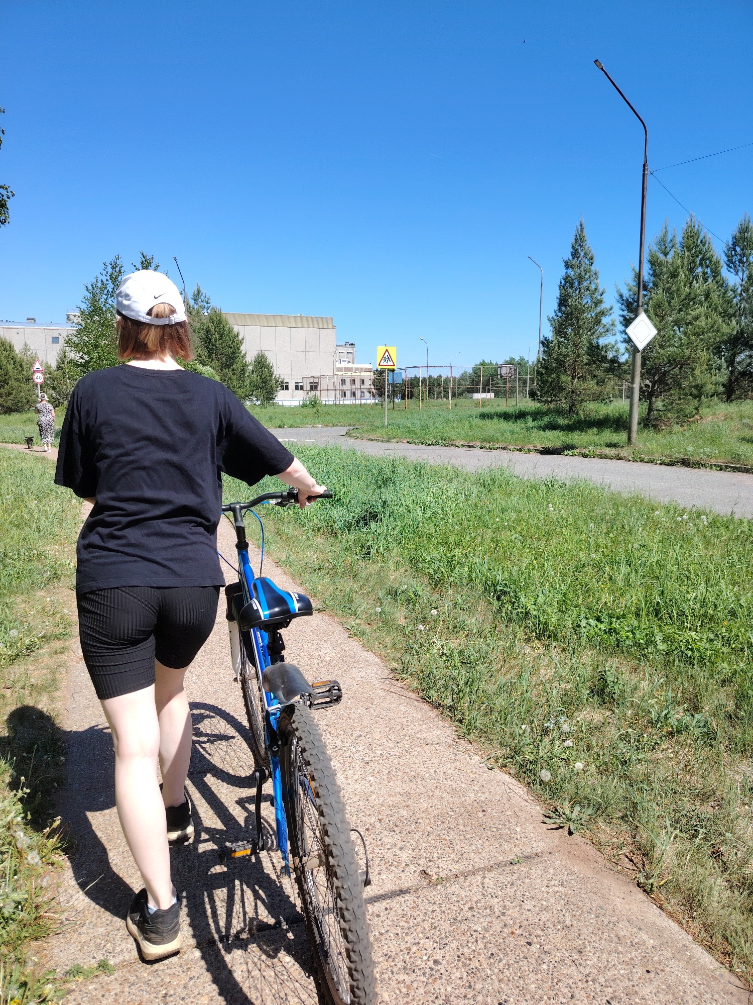 A person is walking along a sidewalk while holding a blue bicycle. The person is wearing a black t-shirt, black shorts, a white cap, and black sneakers. To the right of the sidewalk is a grassy area with trees and signs. In the background, a building and fence can be seen. The sky is blue. The location is Markovsky, Russia.