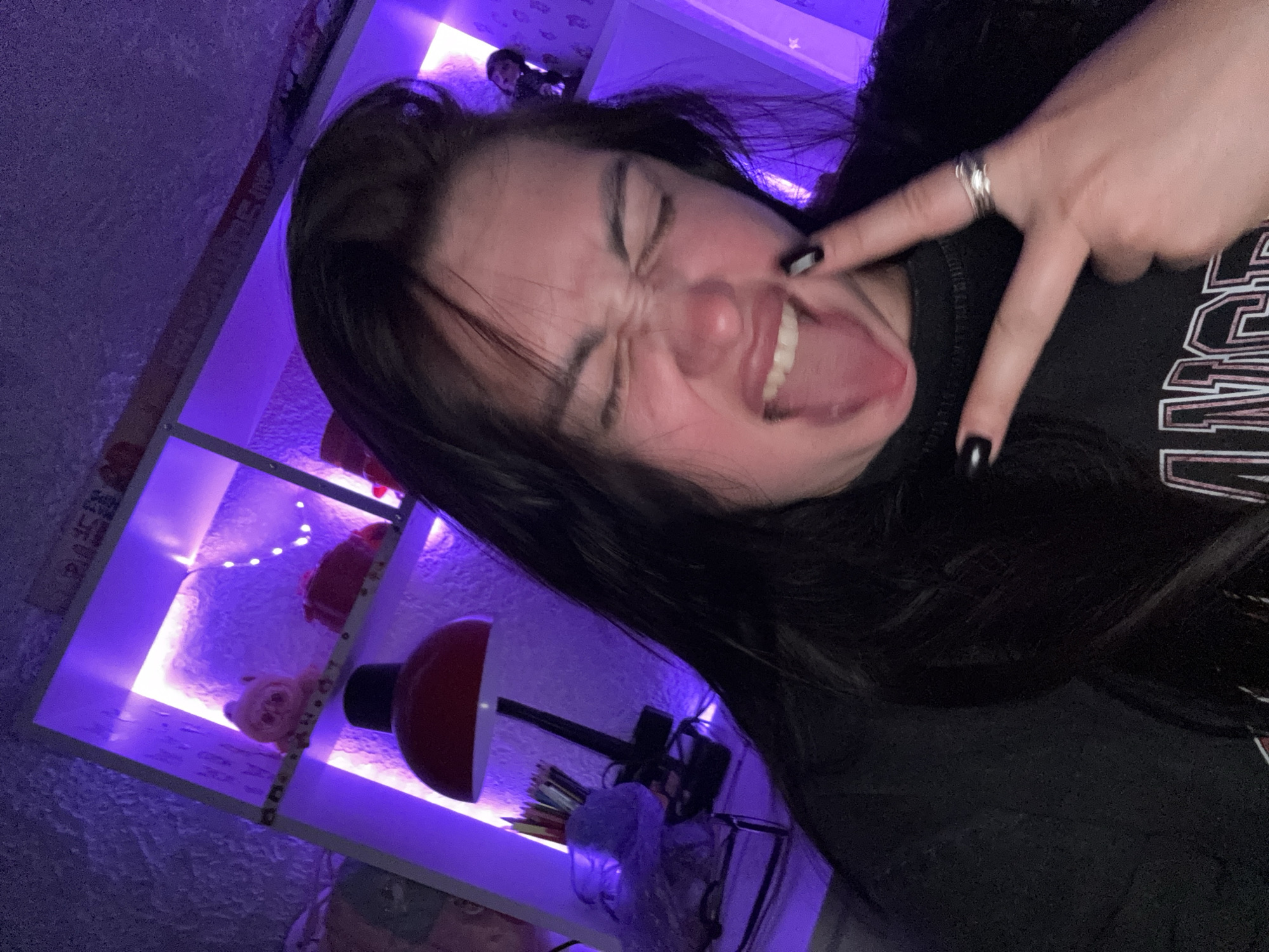 A young person with dark hair and dark nail polish is making a face at the camera, sticking out their tongue and crossing their eyes. They are wearing a black t-shirt with lettering on it. Their right hand is raised, with their index finger and middle finger extended in a peace sign, and they are wearing a ring on their ring finger. In the background, there is a shelf unit illuminated with purple lights. On the shelves, there are various objects including a red lamp, a jar of colored pencils, a small stuffed animal, and what appear to be string lights. The overall environment suggests a bedroom or personal space, lit by artificial purple lighting. No specific location in Tolyatti, Russia is indicated.