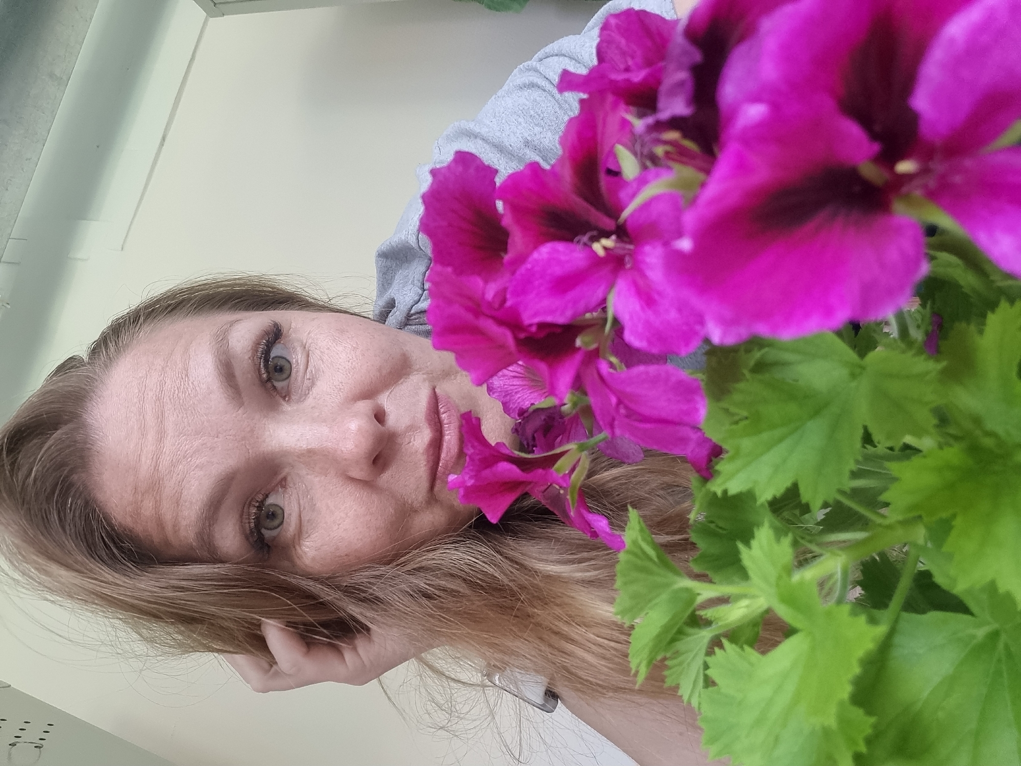 A woman is looking at the camera, with purple flowers in the foreground. The woman has fair skin, light brown hair, and light-colored eyes. She is wearing a gray top. Her hand is near her face. The background is a neutral beige color, possibly a wall. The location is not discernible from the image.