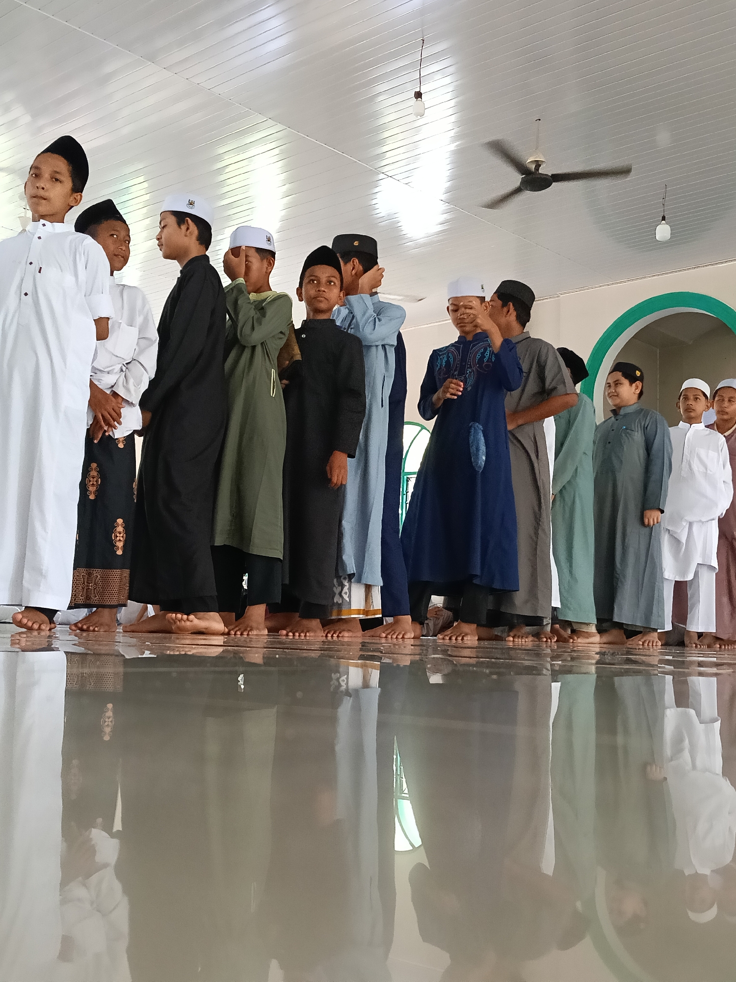 A low-angle shot shows a group of boys standing in a line, reflecting in the polished floor. They appear to be in a religious building in Lhokseumawe, Indonesia. The boys are wearing traditional Muslim attire consisting of long robes in varying colors (white, black, green, blue, grey, and brown) and hats, with some wearing skullcaps. They are barefoot. The building has a white ceiling with a rotating ceiling fan and hanging light bulbs. A green archway is visible in the background.