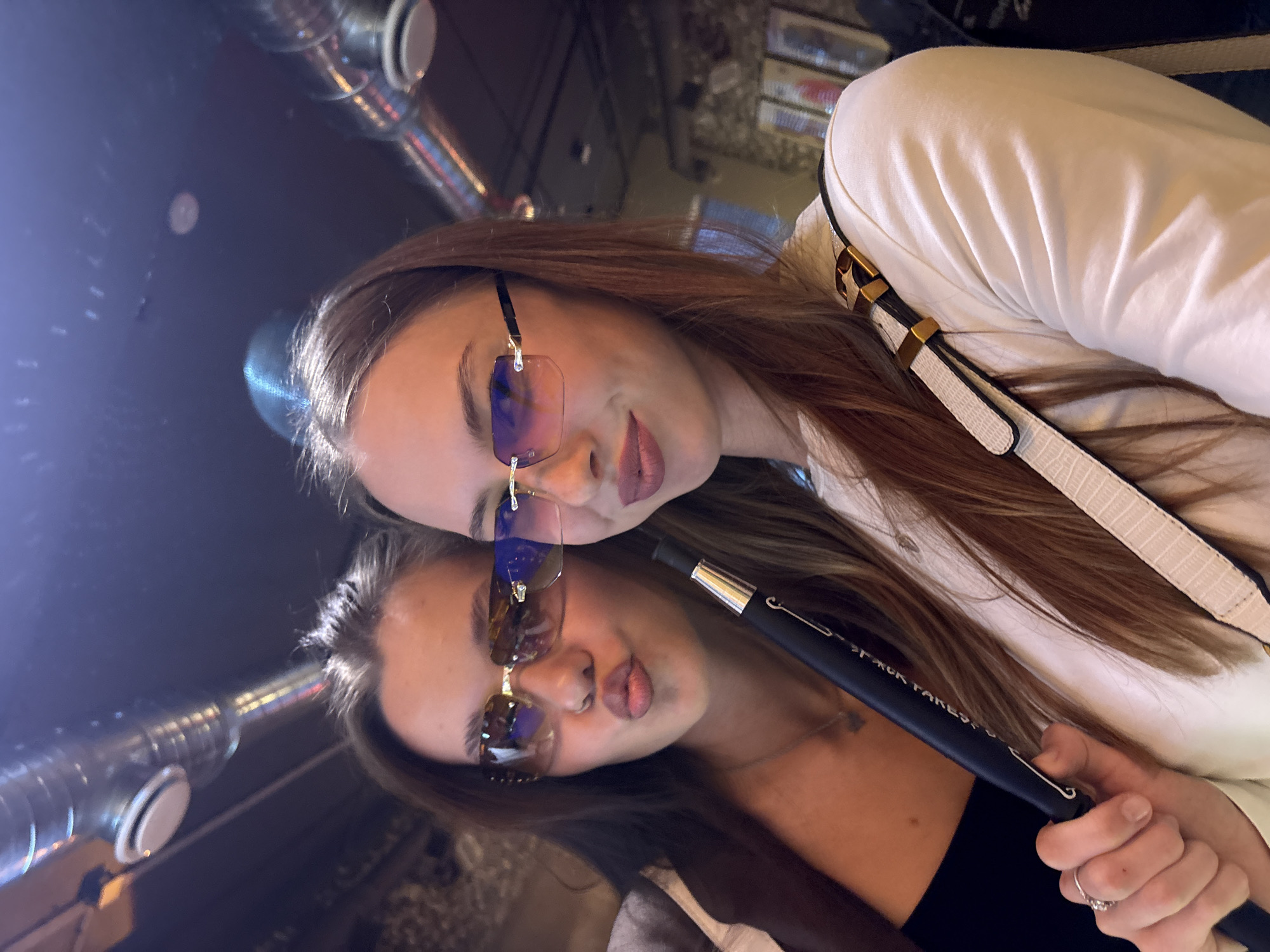 Two young women are taking a selfie indoors. The woman on the left is wearing sunglasses and has her lips pursed. She is holding a black hookah hose. The woman on the right is also wearing sunglasses and smiling. She is wearing a white top and a purse strap is visible over her shoulder. The background appears to be a dimly lit indoor space with exposed pipes on the ceiling. The image does not contain any identifiable landmarks or features that would allow for determination of the specific location within the city of Perm, Russia.