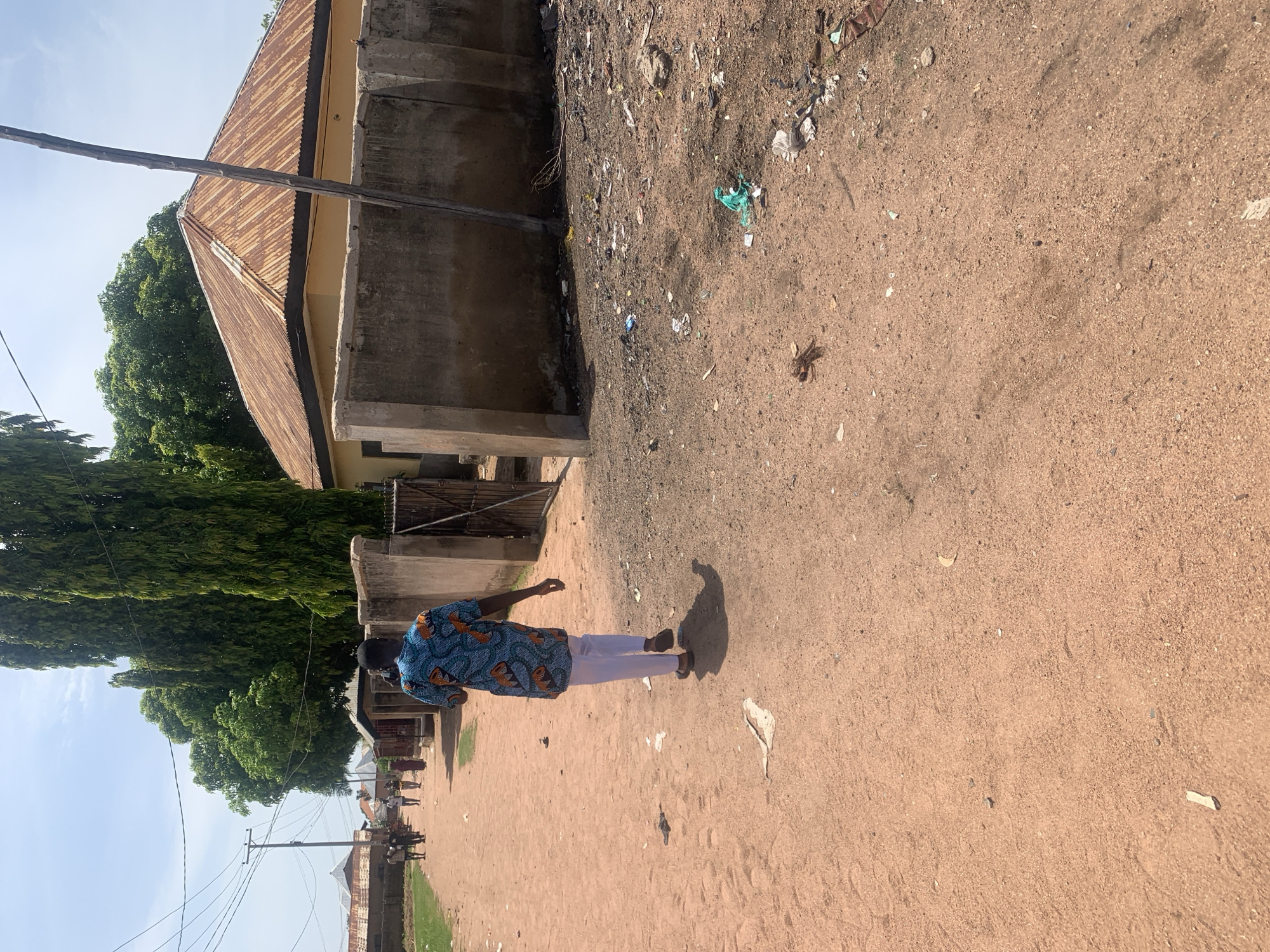 An image captures a dusty, unpaved street in Jalingo, Nigeria, under a bright, hazy sky. On the right side of the street, a light yellow building with a rusty corrugated metal roof stands behind a low concrete wall. Debris and trash are scattered on the ground in front of the wall. Tall, slender trees line the left side of the street, with more buildings and vegetation visible in the background. A lone man, wearing a vibrantly patterned blue and orange shirt and white pants, walks away from the camera down the center of the street. Further up the street, a small group of people are walking in the distance. Power lines are strung across the top of the frame.