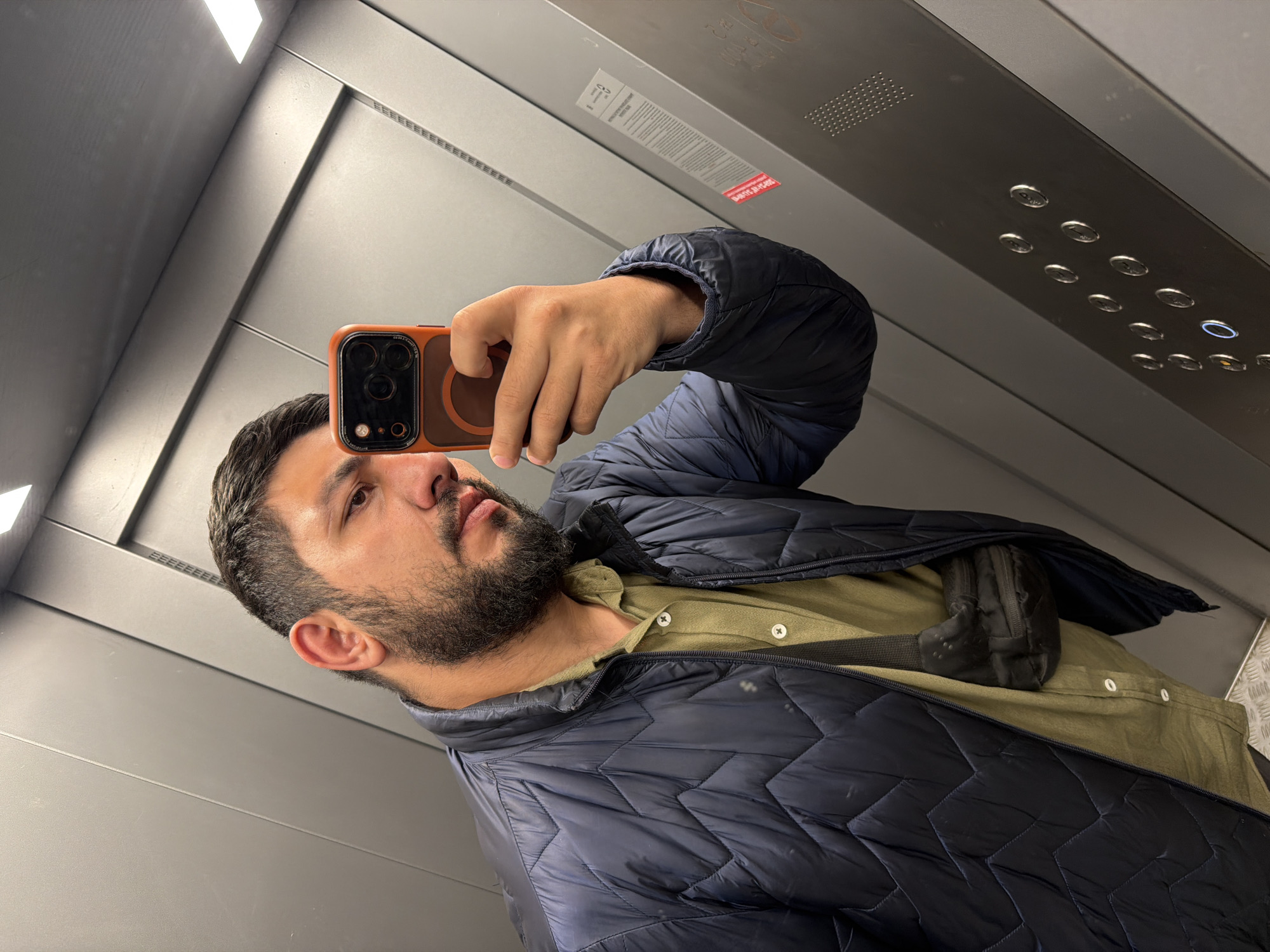 A man with dark hair and a beard takes a selfie in an elevator. He is wearing a blue puffer jacket over a tan button-down shirt and holding an orange-cased phone. The elevator has grey metal walls, a panel with buttons, and a ceiling light. The photograph was taken in the city of Krasnoyarsk, Russia.
