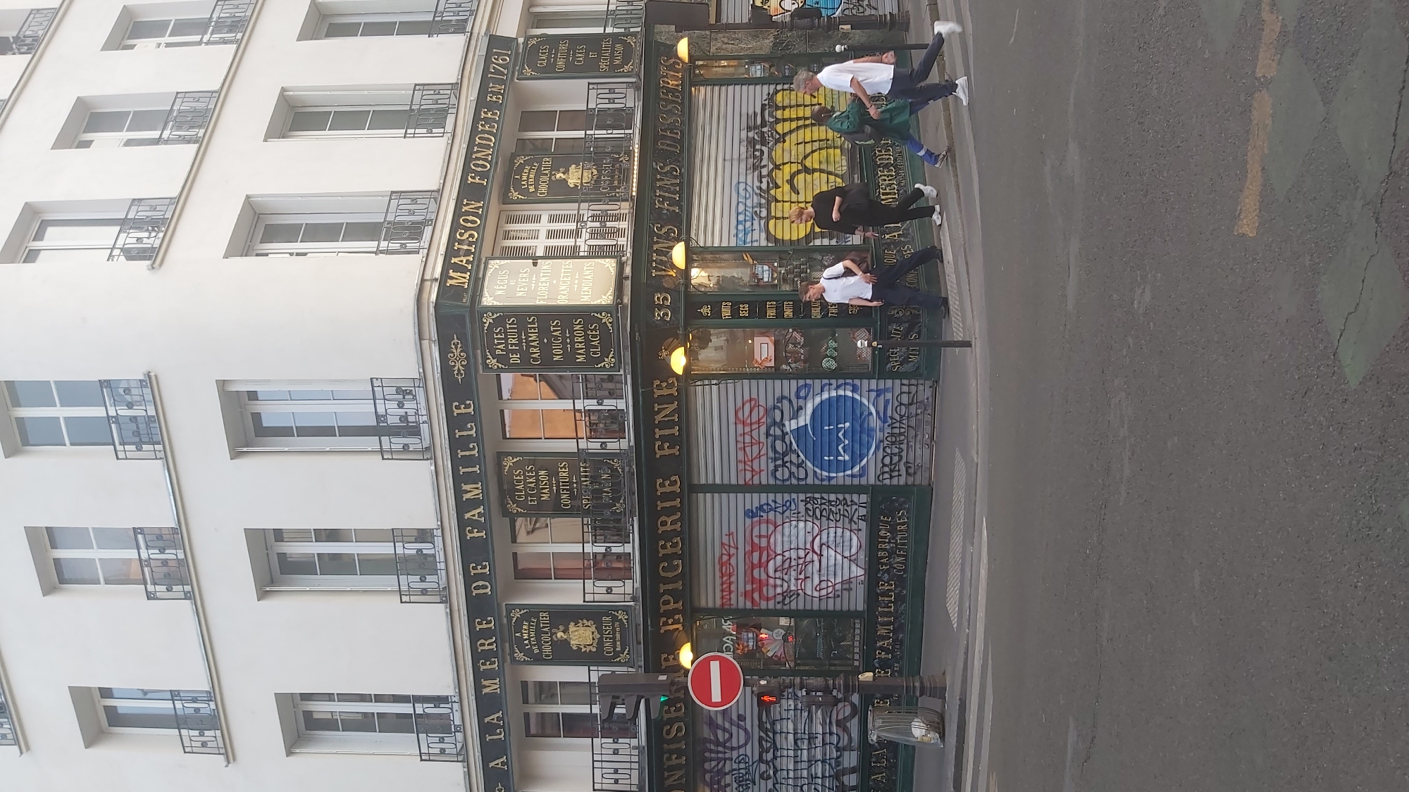 The image depicts a corner storefront in Paris, France, with the text "A LA MERE DE FAMILLE" prominently displayed. The building is white with multiple windows and balconies. The storefront itself has a dark green facade with ornate gold lettering. The lower part of the storefront, including the windows, is covered with graffiti, indicating the shop may be closed or undergoing renovation.
Several individuals are visible walking across the street in front of the store. One person is wearing a white t-shirt and dark pants, another in a black top and pants, a third in a green jacket and blue jeans, and the fourth in a white t-shirt and dark pants. They appear to be pedestrians crossing at an intersection.
Above the storefront, smaller signs list various food items, including "GLACES ET CAKES MAISON" (homemade ice cream and cakes), "CONFITURES" (jams), "PATES DE FRUITS" (fruit jellies), "MARRONS GLACES" (candied chestnuts), and "CHOCOLATIER" (chocolate maker). The text "MAISON FONDEE EN 1761" (House founded in 1761) suggests a long-standing establishment.
A red and white circular stop sign is mounted on a lamppost to the left of the entrance. The street in the foreground is asphalt with painted white and green crosswalk markings. The overall scene captures a street view in Paris, highlighting a historic confectionery and grocery store, albeit with a modern overlay of graffiti on its lower facade.