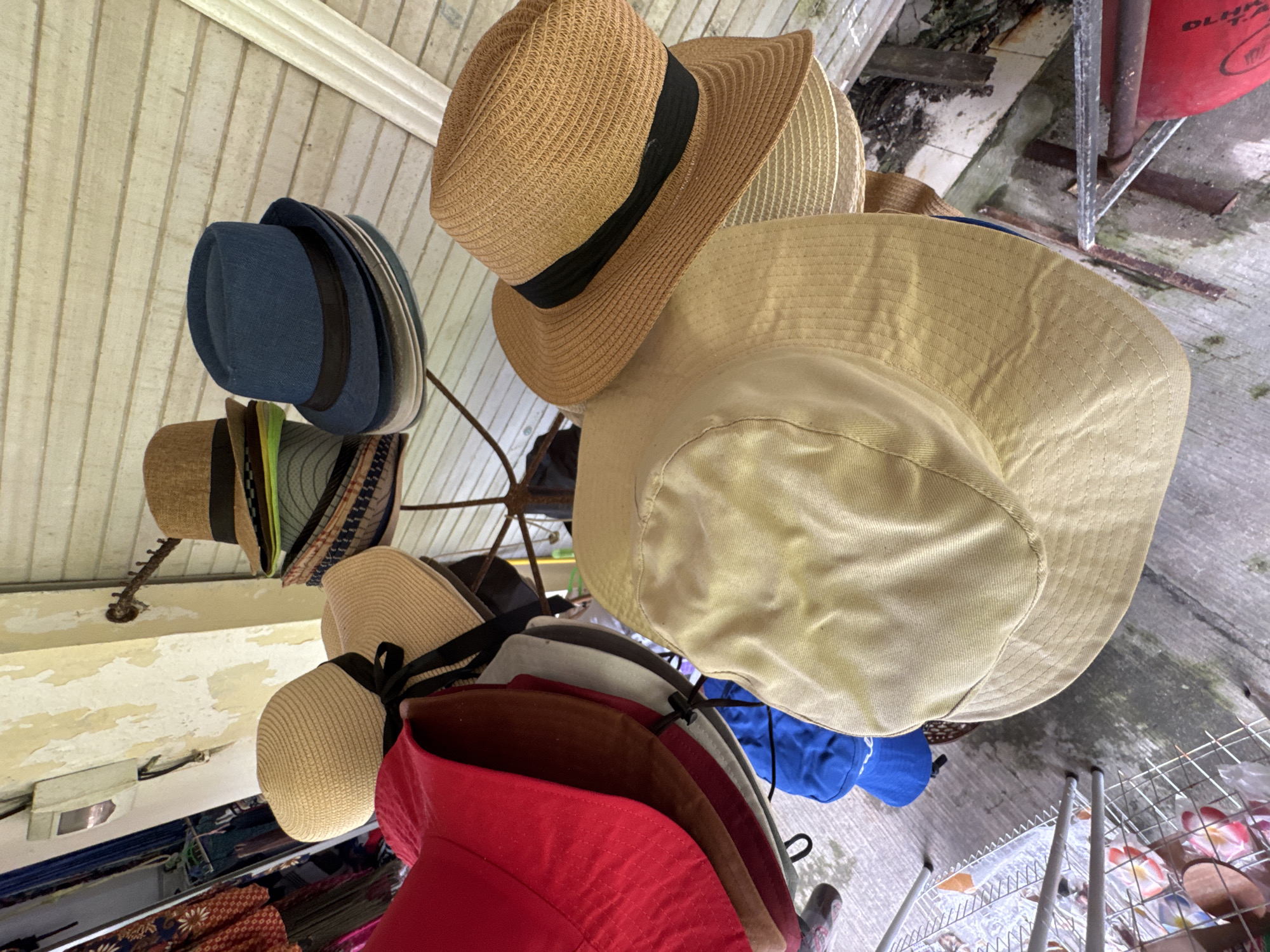 The image depicts a collection of hats on a rack, possibly in a retail setting in Kuta Selatan, Indonesia. The hats vary in style and color, including straw hats, fedoras, and sun hats. A brown straw hat with a black band is prominent at the top. Other hats include blue, red, and beige colors. The background shows a ceiling with white panels and a concrete floor. A wire rack with items is visible in the lower right corner.