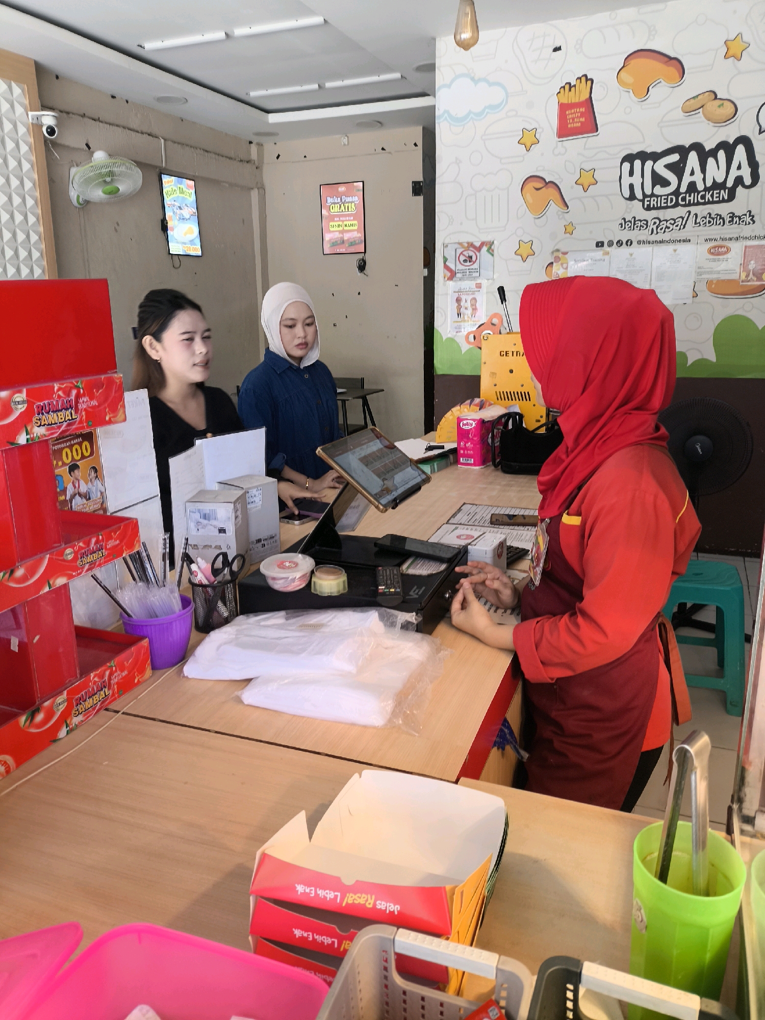 The image captures an indoor scene, likely a fast-food establishment, with a wooden counter dominating the foreground. Two young women are visible, one wearing a black top and the other a blue top and a light-colored hijab. A third individual, wearing a red hijab and a red apron over a maroon shirt, is actively engaged at the counter, possibly a cashier or server. A tablet is positioned on the counter, suggesting a point-of-sale system. Various items are on display and for sale, including red boxes labeled "RUMAH SAMBAL" and what appear to be food packaging or promotional materials. On the wall behind the counter, a large graphic features the "HISANA FRIED CHICKEN" logo and other food-related imagery. A security camera is mounted on the upper left wall. The overall atmosphere suggests a busy service environment. While the location is stated to be Gorontalo, Indonesia, there are no specific visual cues within the image that definitively confirm this precise geographical location.