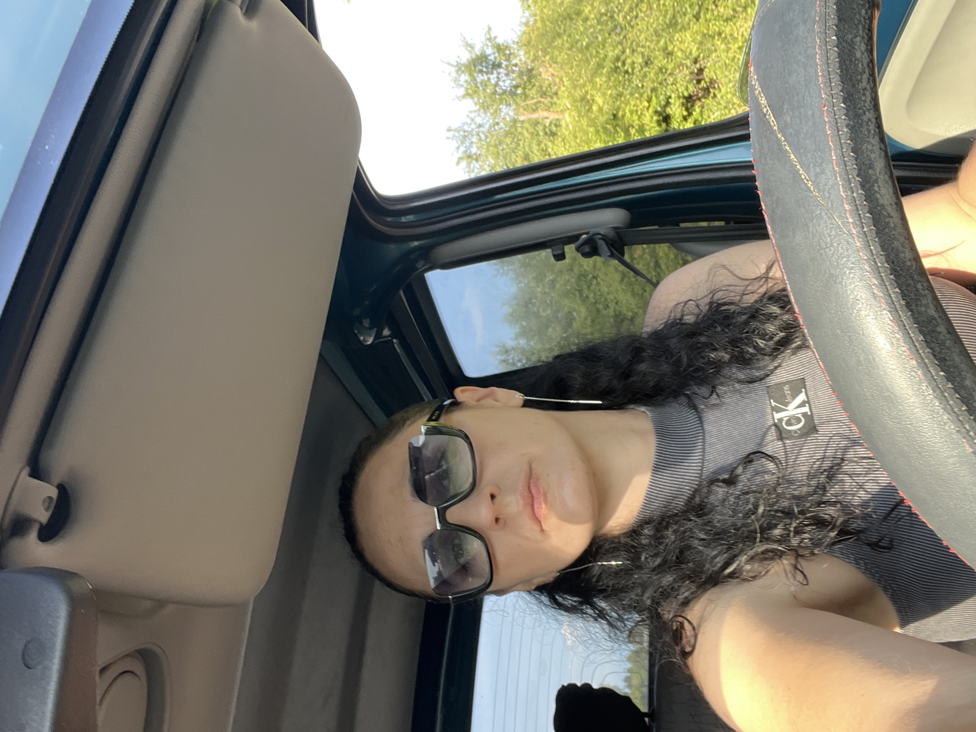 A medium shot photograph shows a woman driving a car in Tarnivtsi, Ukraine. The woman, with dark, curly hair, wears sunglasses and a grey tank top with a "CK Jeans" logo. She is looking forward, with her hands on the steering wheel. The car interior is visible, including the sun visor and part of the dashboard. Outside the car, lush green trees and vegetation are visible, suggesting a rural or suburban setting. The sunlight casts shadows, indicating it is daytime.