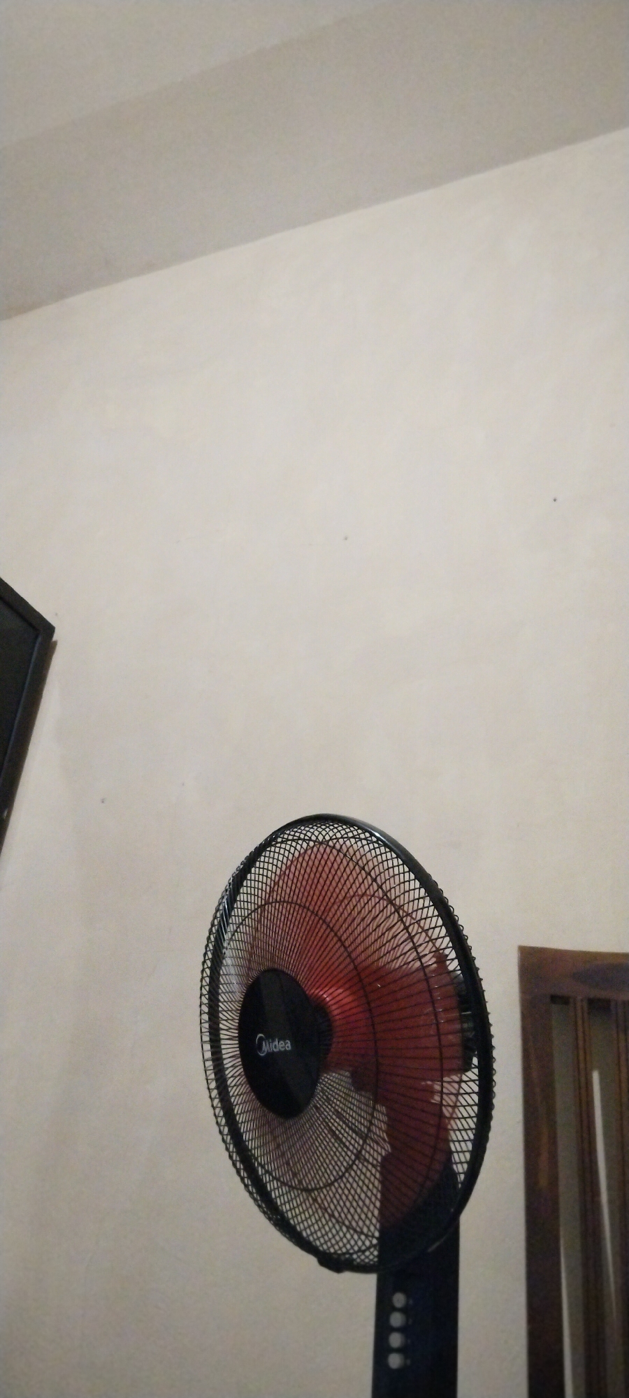 This media file shows a close-up of a black and red oscillating fan in an indoor setting. The fan is positioned in the lower right quadrant of the frame, with its blades blurred due to motion, suggesting it is in operation. The fan's guard is black mesh, and the blades are a vibrant red. A black circular emblem with the text "Midea" is visible on the front of the fan's motor housing. The fan's stand is black and extends downwards out of frame, with several circular buttons visible near the top.
The background is a plain, light-colored wall, likely painted. In the upper left corner, a portion of a dark object, possibly a television screen, is visible. To the right of the fan, part of a wooden piece of furniture, likely a cabinet or chair, with vertical slats can be seen. The lighting appears to be artificial and somewhat dim. The overall scene suggests a domestic or office environment. No people are visible in the media file. The time of day and weather conditions are not discernible from the image. The location is noted as Duman, Indonesia.