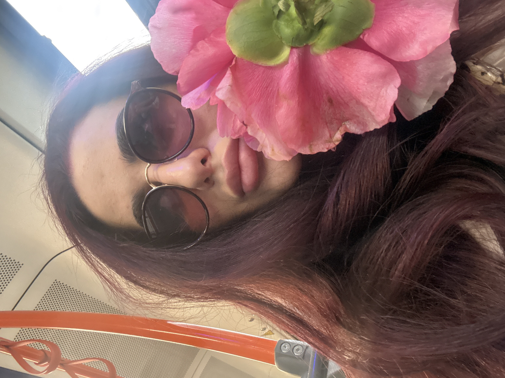 A selfie-style photograph taken from a low angle features a person with long, reddish-brown hair. A large pink flower with a green center is positioned as a hair accessory, partially obscuring the person's forehead and right eye. The person is wearing round sunglasses with a gold nose ring. The background appears to be the interior of a public transportation vehicle, possibly a subway or bus, as suggested by the overhead lighting and patterned paneling. The presence of an orange rail suggests that the vehicle is a Sofia public transport train.