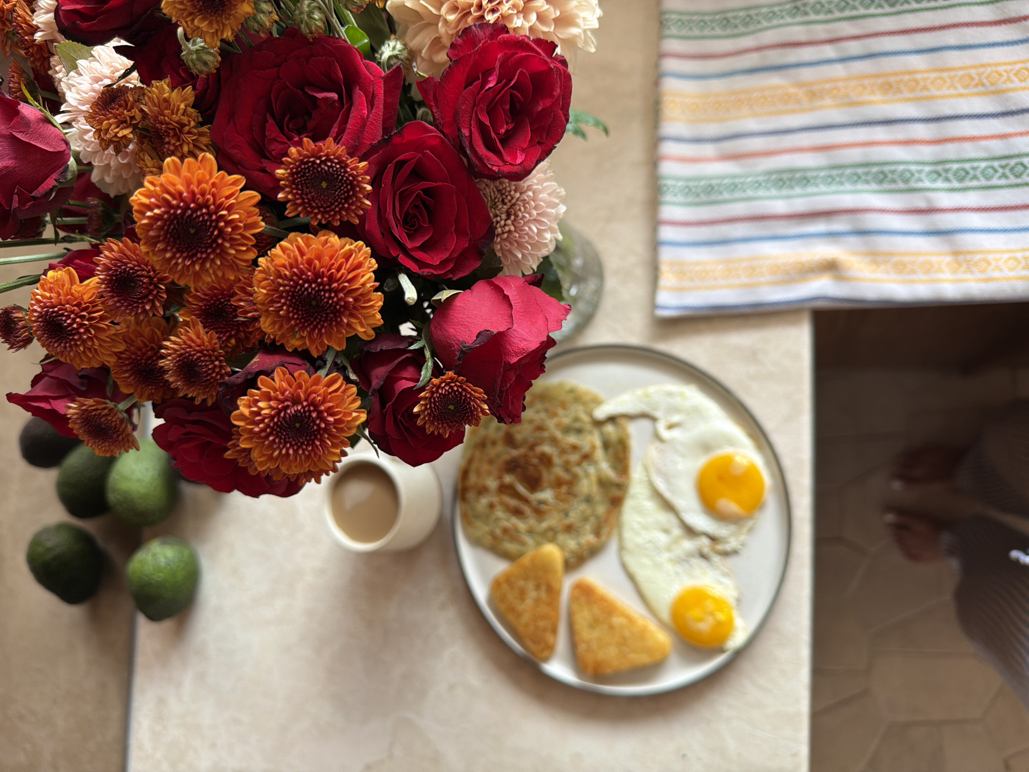 The image depicts a breakfast scene on a table. A plate holds two fried eggs, a flatbread or pancake, and two triangular hash browns. Beside the plate is a white mug of coffee. A vase of red and orange flowers sits next to the coffee, with several green limes placed to the left of the vase. A striped towel is visible in the upper right corner. In the extreme lower right of the image, a person's feet and lower legs are visible. The table surface appears to be beige with a subtle pattern. The location, Mengwi, Indonesia, is provided, though not visually confirmed in the scene.