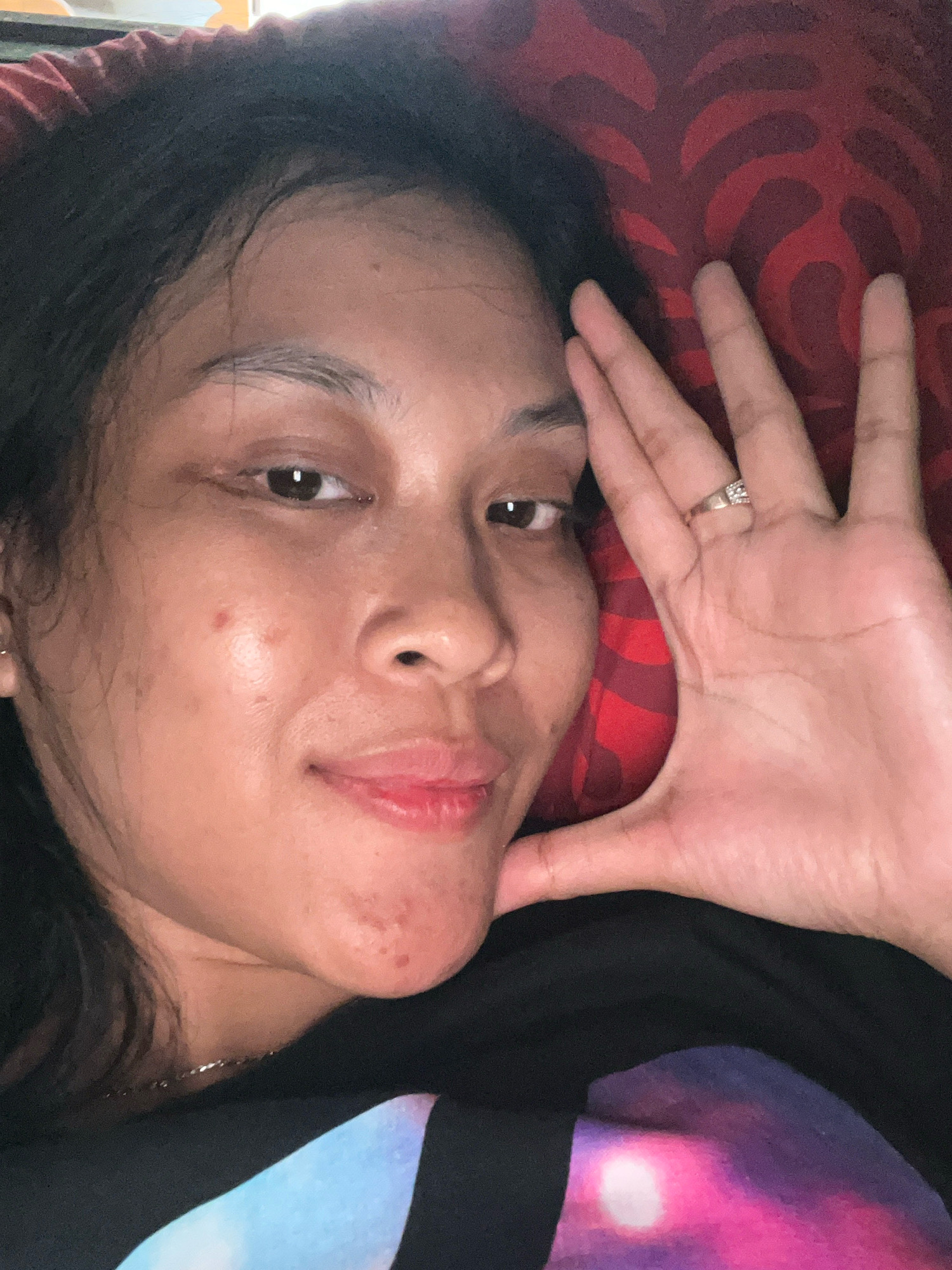 The image is a close-up selfie of a woman lying on a red patterned pillow. She has dark hair, dark eyes, and fair skin. She is wearing a black t-shirt with a colorful graphic design. Her right hand is raised to her face, with a ring on her left ring finger. The image appears to be taken indoors, possibly in a bedroom, in Kecamatan Pati, Indonesia.