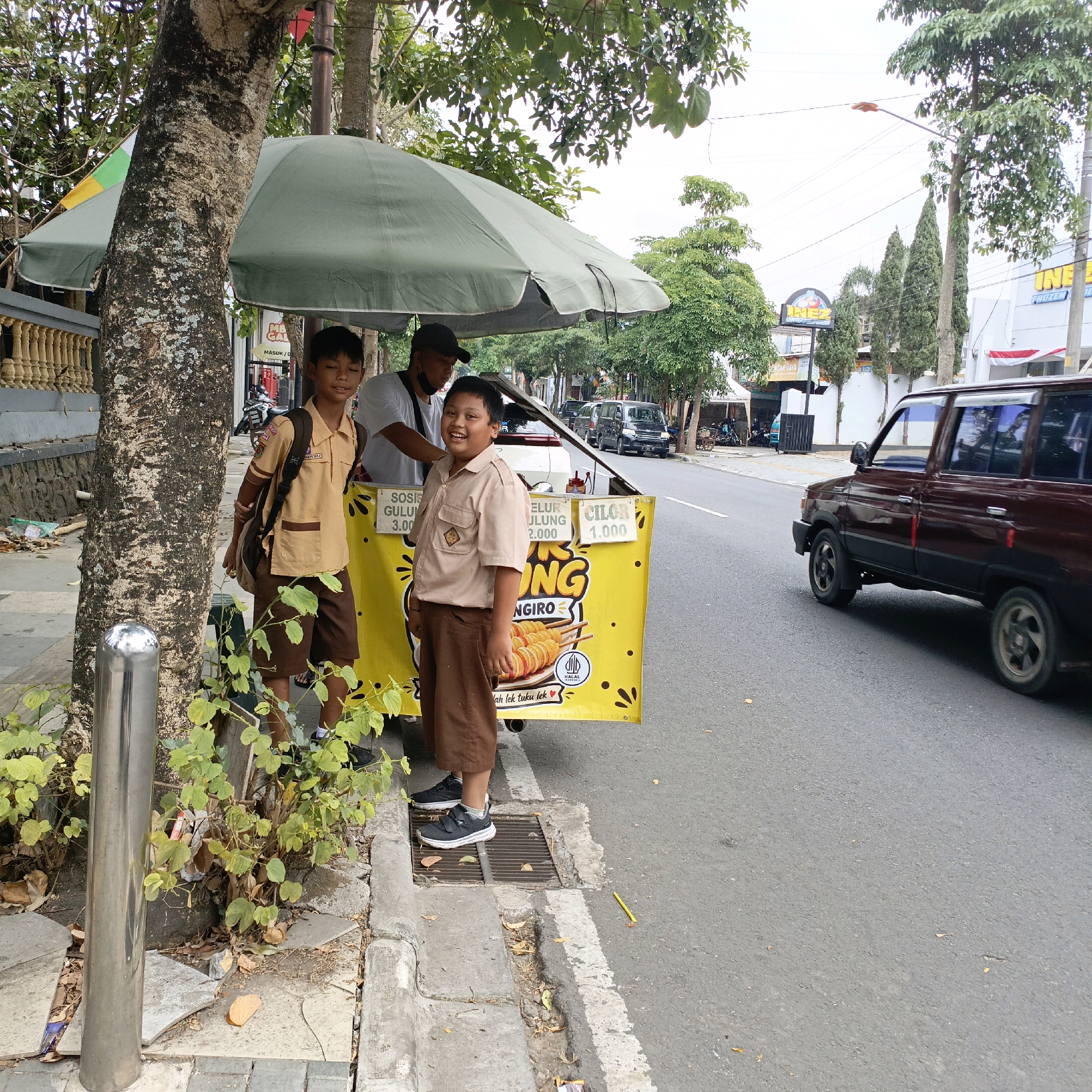 The image depicts a street scene in Salatiga, Indonesia, where a food cart is set up under a large green umbrella. Two young boys in school uniforms, a tan shirt with a badge and brown pants, stand near the cart. One boy is looking at the camera with a smile, while the other is partially visible to his left. A man wearing a black cap and a white t-shirt stands behind the cart, appearing to be serving food. The food cart has a yellow banner with text and images of food items. The cart is positioned on the sidewalk next to a busy road. A maroon minivan is driving on the road to the right of the cart, and other vehicles are visible in the background. Trees and buildings line the street. The sidewalk is made of concrete and pavers, with a drainage grate visible. There is some greenery growing on the side of the sidewalk. The overall scene suggests a typical day in a bustling urban area of Indonesia.
