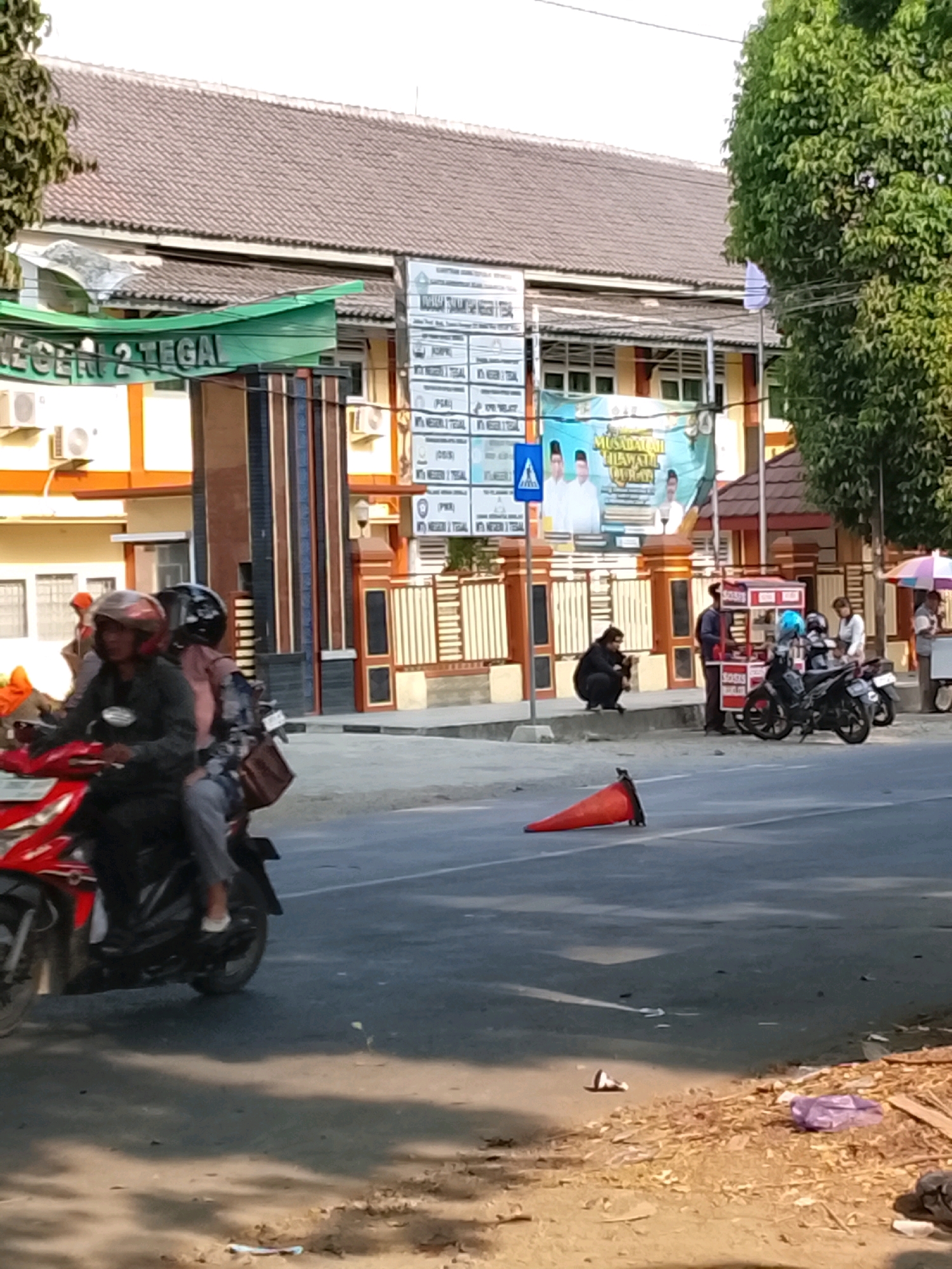 The image captures a street scene in front of a building that appears to be a school or public institution, likely located in Slawi, Indonesia, given the context. The building has a tiled roof and a banner that reads "LEGER 2 TEGAL". A bright green awning extends over part of the entrance. In front of the building, there is a sidewalk with several people and some vendors. One person is sitting on the curb, looking at their phone. Two motorcycles are parked, one with a blue helmet on it. Further down the sidewalk, a food stall is visible, with people gathered around it. A person is holding an umbrella. On the street, a red motorcycle with two riders is in motion, heading towards the viewer. A traffic cone is lying on its side in the road. The ground is paved, and there is some debris on the side of the road, including wood scraps and a purple plastic bag. The overall scene is consistent with a typical urban streetscape in Indonesia.