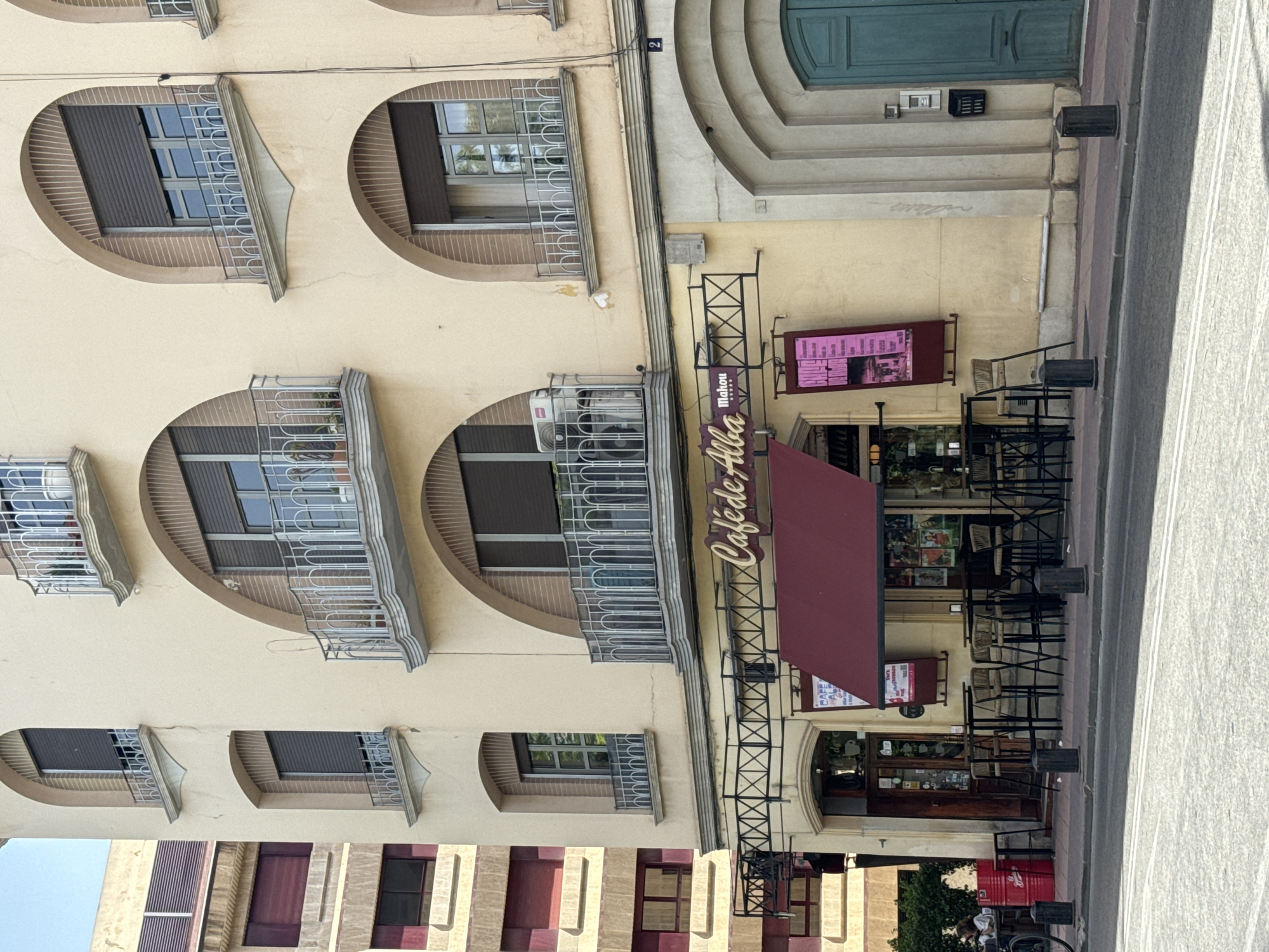 This is an outdoor, eye-level, medium shot of the facade of a building in Murcia, Spain. The building's facade is painted a pale yellow and features several arched windows with metal grates. Some of the windows have dark-colored shutters. A section of the building has a burgundy awning extended over an entrance. Above the awning, a sign reads "Café de Alba." There are tables and chairs set up outside the cafe, some of which are occupied by customers. The scene appears to be a sunny day, and the overall atmosphere is casual and relaxed.
Further down the street, there are other buildings with similar architectural features. The image also captures a street with a sidewalk and a road. A red recycling bin is visible near the foreground, and a person is seen sitting on a stool near the bin. The overall impression is a typical urban street scene in Spain on a bright day.