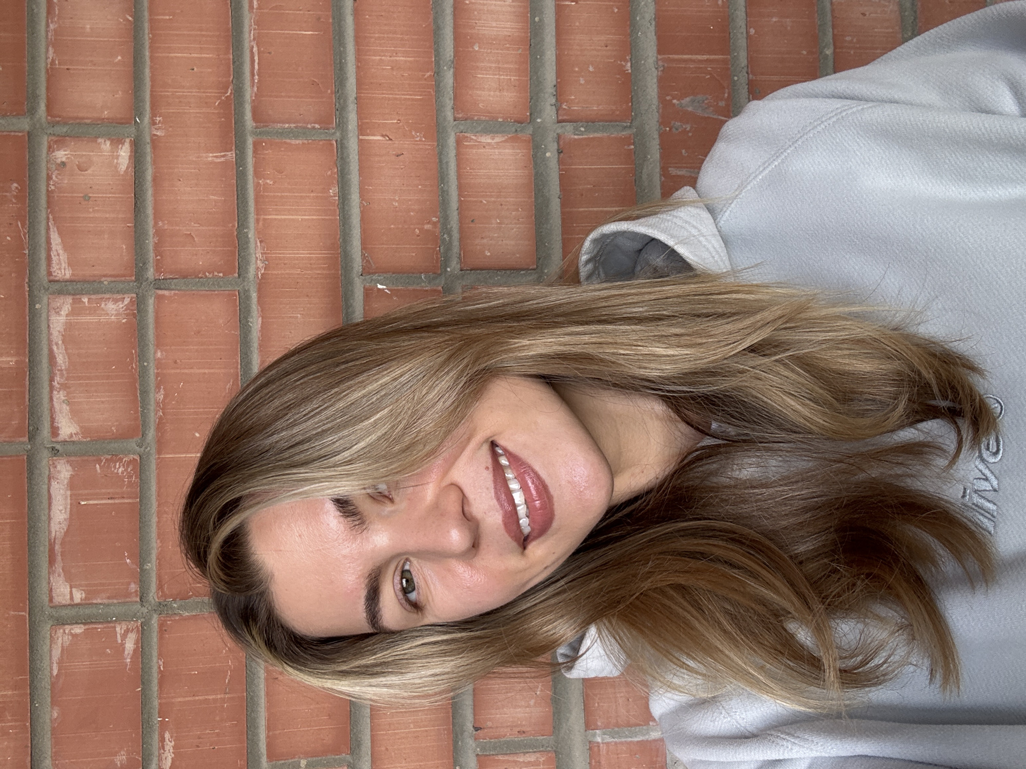 A young woman with long, wavy, blonde and light brown hair is depicted in a close-up portrait against a reddish-brown brick wall. Her head is tilted, and she is smiling broadly, revealing her teeth. She has hazel or green eyes, with one eye clearly visible and looking towards the viewer. Her lips are adorned with a pinkish-nude lipstick. She is wearing a light gray hooded sweatshirt (hoodie), with some stitching visible on the shoulder. Subtle, embroidered text, partially obscured by her hair, is visible on the right side of her sweatshirt, appearing to spell "COLLEGE". The brick wall background features rectangular bricks with visible gray mortar lines, showing some texture and light discoloration on the brick surfaces. The overall lighting suggests daylight.