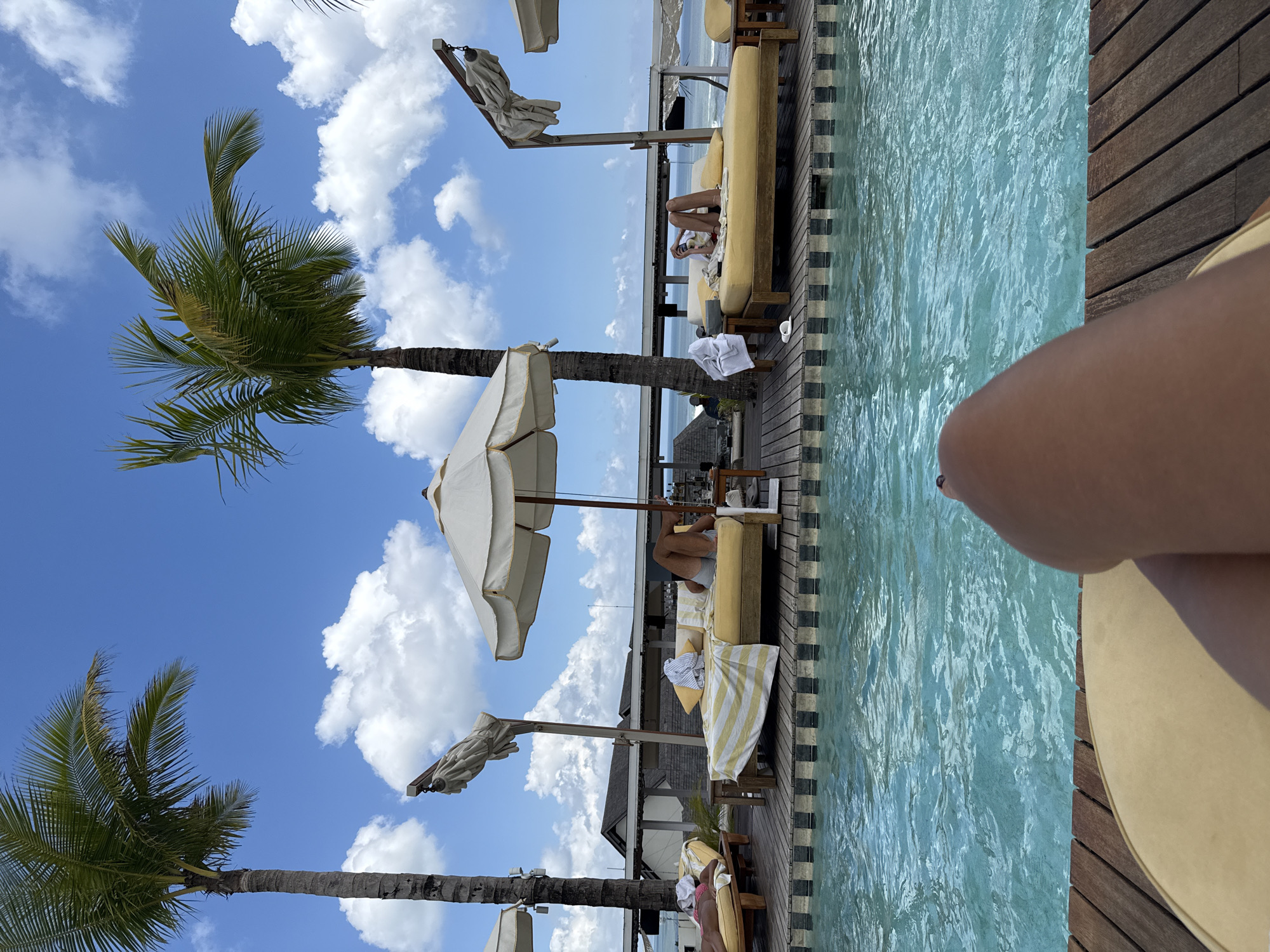 The image captures a poolside scene in North Kuta, Indonesia, under a partially cloudy blue sky. Two palm trees frame the left side of the image. A person is relaxing on a yellow lounge chair next to a pool, with part of their leg visible in the foreground. Two additional yellow lounge chairs are visible in the background, with individuals reclining on them. Each lounge chair is shaded by a white parasol and has white towels. A multi-level building is also visible in the background. The pool is bordered by a wooden deck with black and white patterned tiles on one side and plain wooden planks on the other. The water in the pool has a light blue tint.