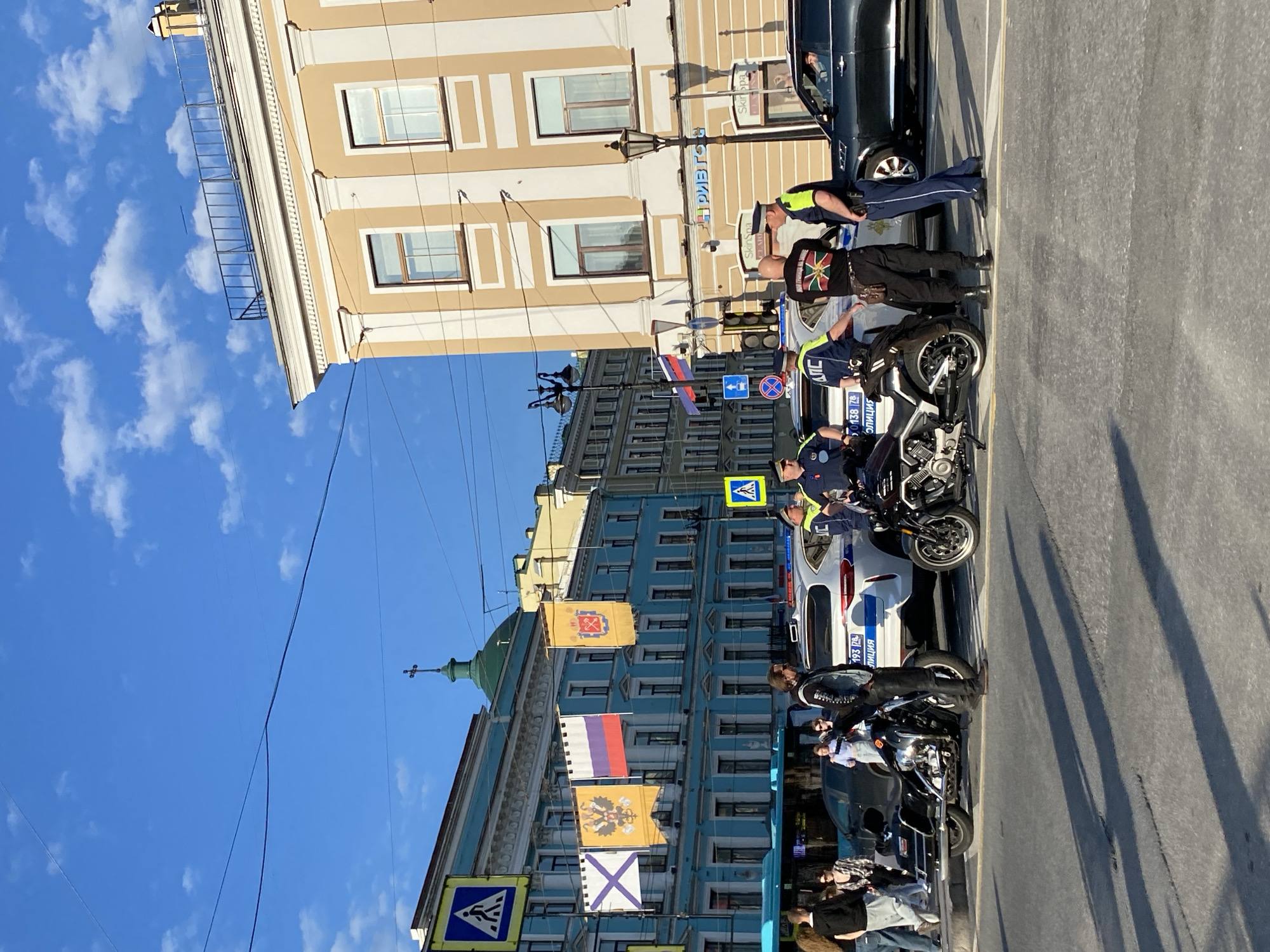 This image captures a vibrant street scene in Saint Petersburg, Russia, under a clear blue sky with scattered white clouds. The bright daylight creates long shadows on the asphalt road, suggesting it is late morning or early afternoon.
The central focus is a gathering of several police officers and their vehicles. At least two police motorcycles, dark in color, are parked on the side of the road. A white police car, marked with "ПОЛИЦИЯ" (POLITSIYA - Police) and the number "193" on its side, is also present. Several police officers are standing around the vehicles; some wear standard dark blue uniforms with light blue/white shirts, while others have reflective yellow vests. One officer is seen interacting near a dark SUV in the background. Among the police, a civilian is visible wearing a black t-shirt with a distinctive circular emblem featuring a lion or sunburst pattern in vibrant colors. Other civilians are partially seen in the background.
The street is lined with historic buildings. On the right, a large, light tan building with white trim and numerous windows stands prominently. A sign on this building partially shows "ПРИВЕТ" (PRIVET - Hello) and another, "Skiripa." On the left, a more elaborate blue building features a green dome topped with a cross, indicative of a church or historic structure. From this blue building, several flags are displayed: a golden flag with a red emblem, a Russian tricolor flag (white, blue, red), a golden flag with a black double-headed eagle (Imperial Russian standard), and a white flag with a blue saltire (St. Andrew's flag). Streetlights, traffic lights, and pedestrian crossing signs are also visible, contributing to the urban environment. The scene appears calm and orderly, suggesting a routine police presence.