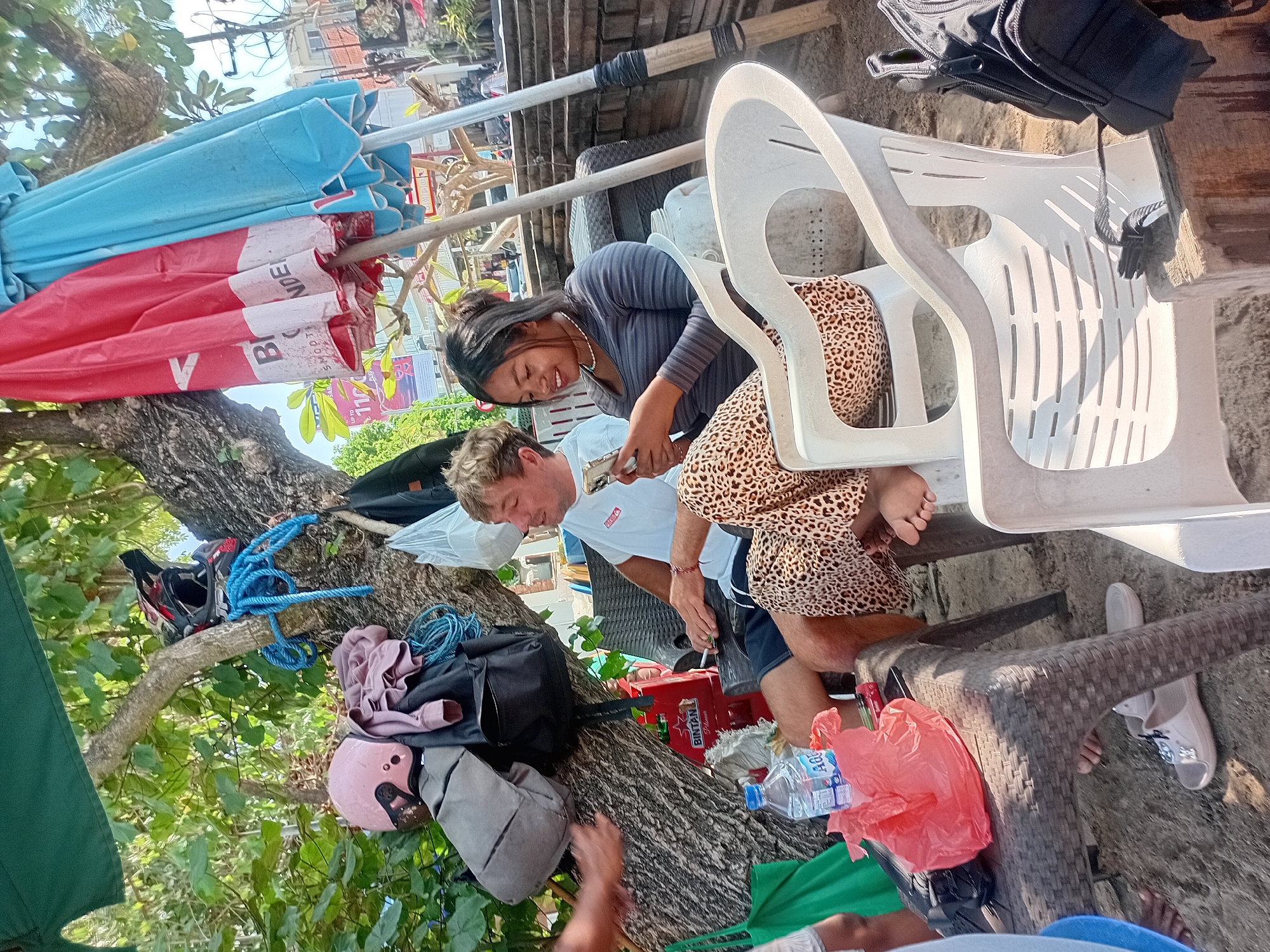 The image depicts a scene on a beach in Kuta, Indonesia, likely a rest stop or seating area under trees. In the foreground, two individuals are seated in white plastic chairs. A man with blonde hair, wearing a white t-shirt and blue shorts, is seated on the left, holding a smartphone and a cigarette. To his right, a woman with dark hair, wearing a grey long-sleeved shirt and leopard-print pants, is also looking at her phone.
In the background, a tree trunk displays several items: a red and black motocross helmet, a pink scooter helmet, backpacks, and coiled blue rope. Two large beach umbrellas, one red and one blue, are propped up. A low table sits between the chairs, holding a bottle of "AQUA" water, a red plastic bag, and a red crate with "BINTAN Premium" visible. The ground appears to be sandy, consistent with a beach environment. Other elements in the background suggest a developed area with structures and possibly other people. The lighting indicates it is daytime, with sunlight filtering through the tree canopy.