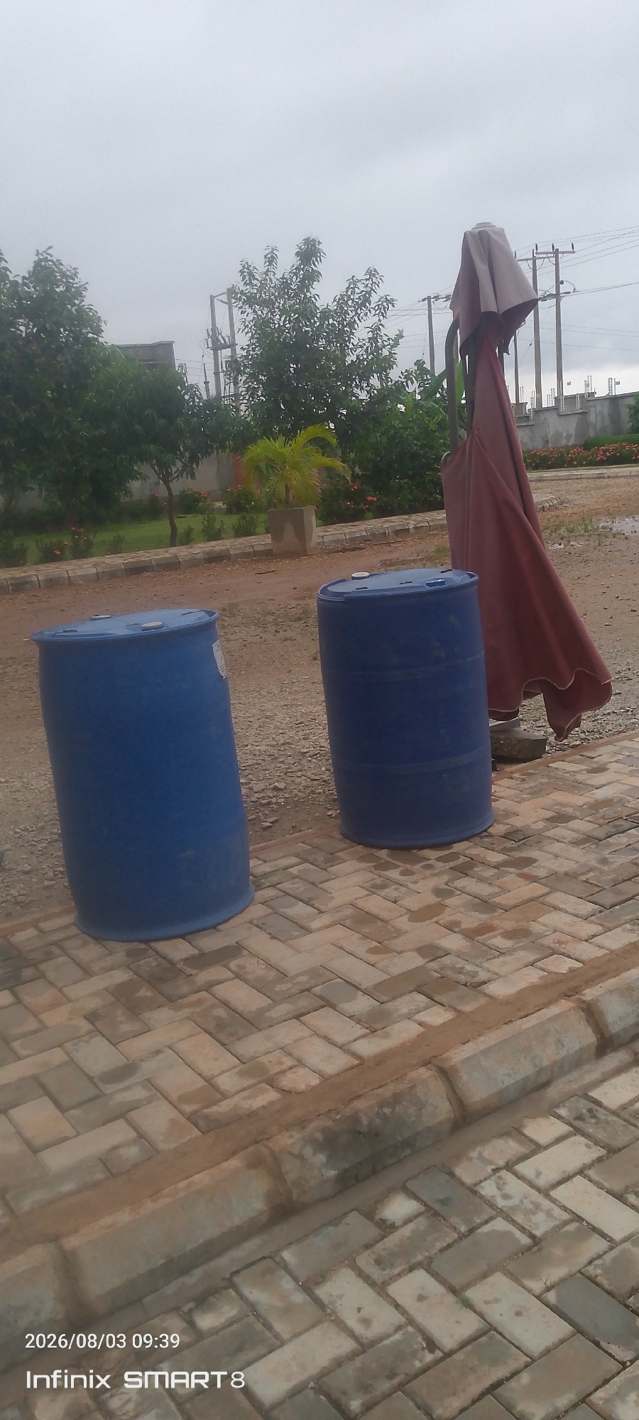 The image depicts an outdoor scene in Kakau, Nigeria, on a cloudy day. In the foreground, two large blue plastic barrels are visible, placed on a cobblestone pathway. One barrel is to the left, and the other is to the right, slightly further back. To the right of the barrels, a large, red umbrella is partially furled and leaning on a stand. Behind the barrels and umbrella, a patch of dirt and gravel is visible. Further in the background, there is a green lawn area with trees and a potted plant. A concrete wall and utility poles with electrical wires are also present in the background. The overall environment suggests an outdoor area, possibly a yard or a public space. The timestamp on the image indicates it was taken on August 3, 2026, at 09:39.