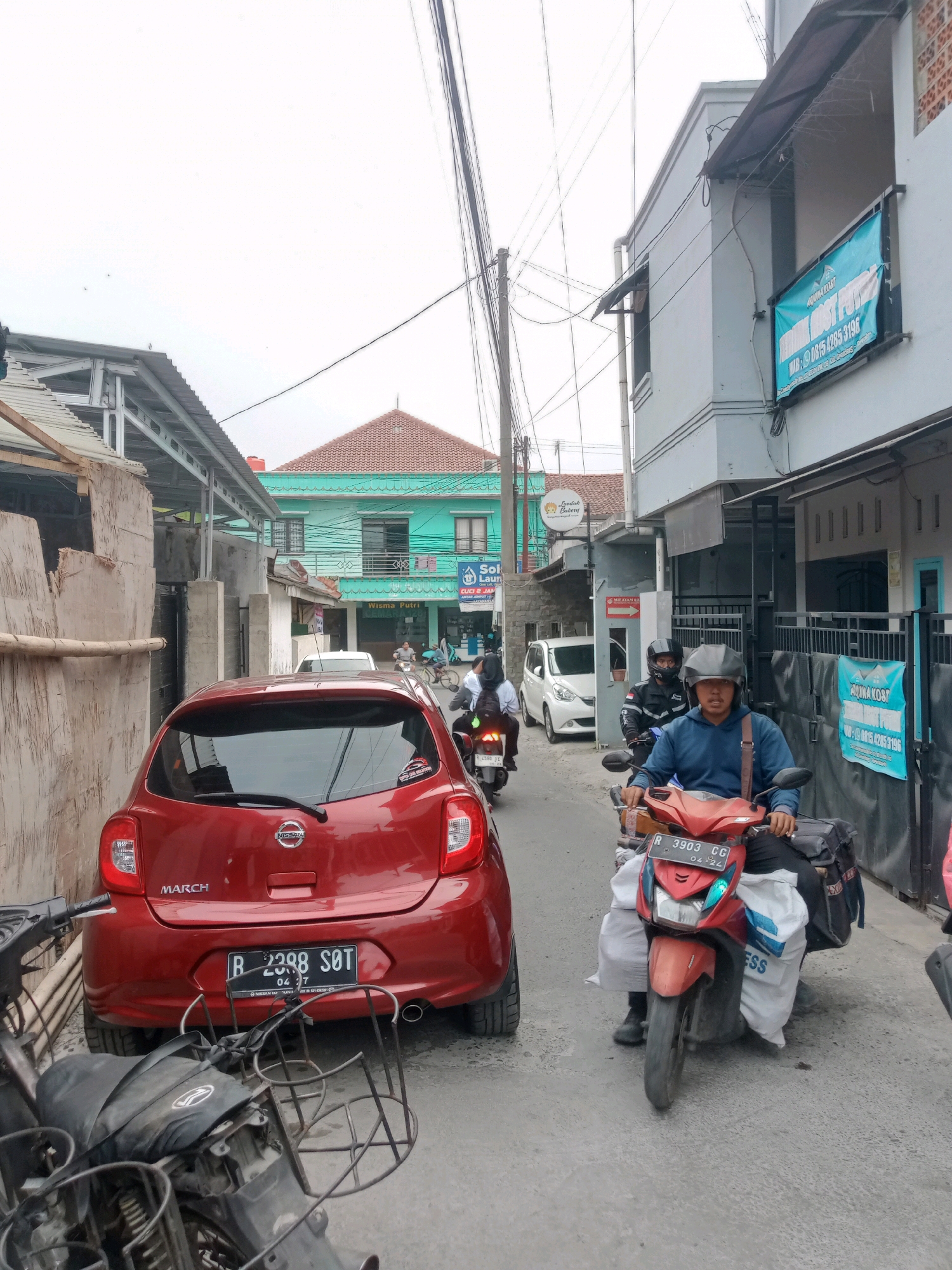 The image depicts a street scene in Grendeng, Indonesia, with a red Nissan March car in the foreground. A man wearing a blue jacket and a helmet is riding a red motorcycle, carrying large white bags on either side of the vehicle. He is looking towards the camera. Behind him, a white car is visible, and further down the street, more vehicles and people can be seen, including a cyclist and another person on a motorcycle. Buildings line the street, some with shops displaying signs, including one for "Wisma Putri." The sky is overcast. The overall scene suggests a busy urban environment.