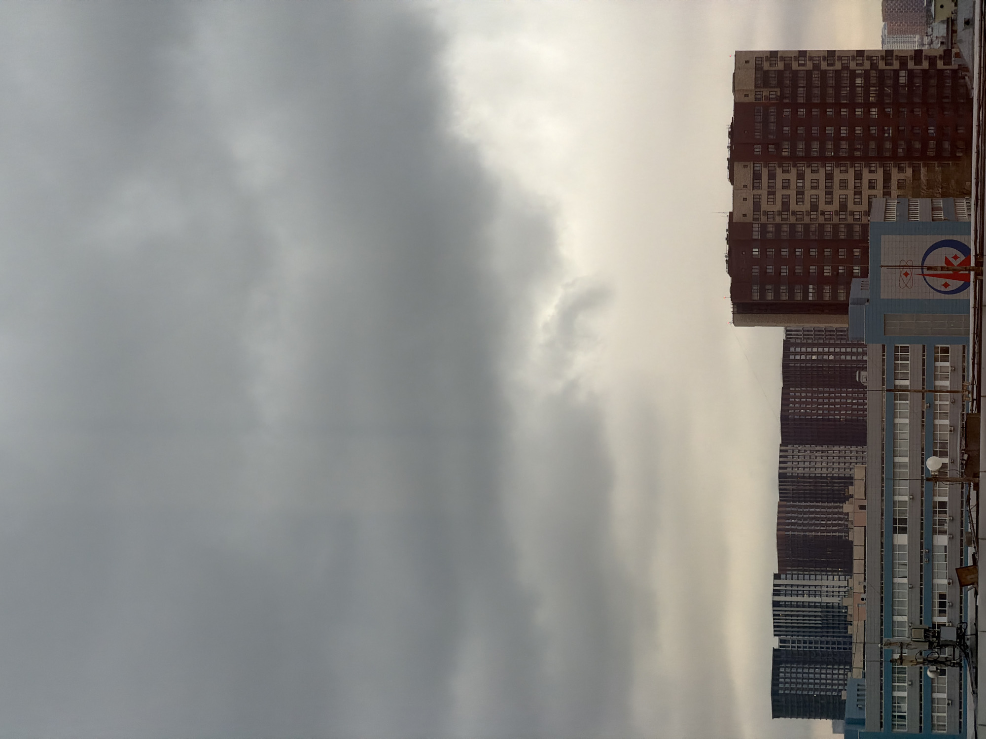 The image shows a cityscape in Moscow, Russia, under a cloudy sky. A row of tall residential buildings lines the horizon, with varying colors and architectural styles. The sky is overcast, with shades of gray dominating the upper portion of the frame. A building with an Atomenergomash logo is visible. The photo appears to be taken from an elevated position.