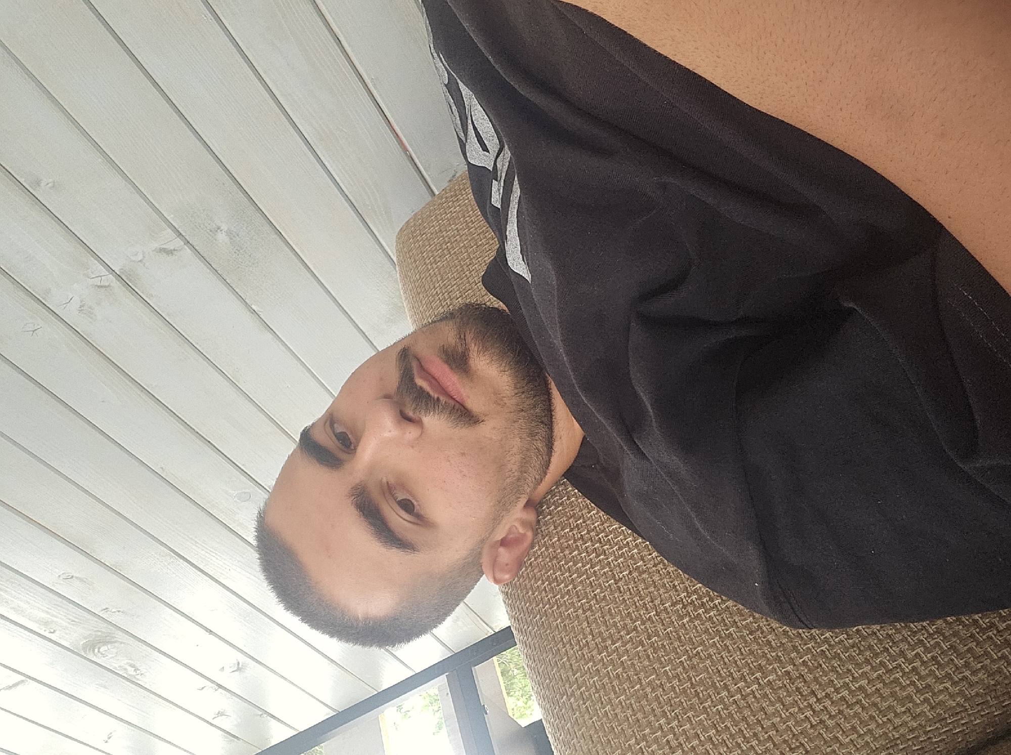 A close-up, over-the-shoulder selfie is taken from a low angle of a young man with short dark hair and a beard, resting on a textured couch. He is wearing a black t-shirt with a grey graphic design on the sleeve. The background consists of a light-colored, slatted wooden ceiling, and a partial view of outdoor greenery through a window on the left. The image appears to have been taken during daylight hours, possibly on a patio or porch.