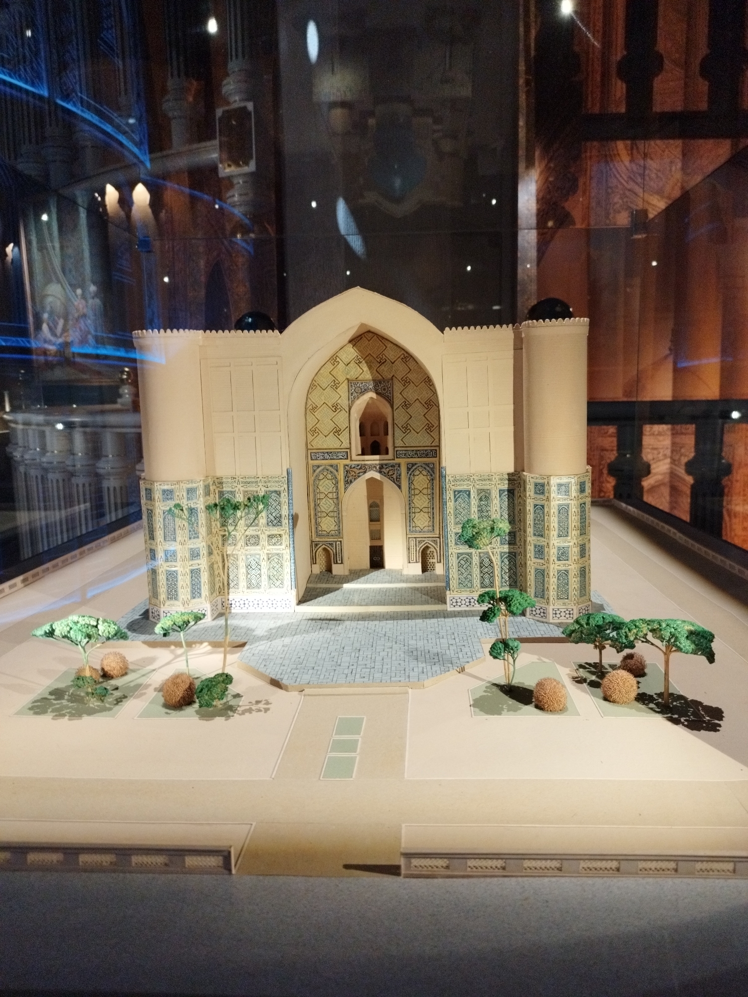 The image shows a detailed architectural model of a building with Islamic influences, possibly a mosque or mausoleum, displayed within a museum setting. The model features a central arched entrance with intricate blue and white tile work, flanked by two cylindrical towers. The facade of the building is adorned with decorative panels and patterned tiles. In front of the structure, a paved courtyard is laid out, with a miniature garden area containing model trees and bushes. The entire model is presented on a light-colored base, with a reflective glass enclosure in the foreground. The background of the image shows the museum environment, with reflections of interior architecture and lighting, including decorative columns and glowing lights, suggesting it is part of an exhibition. The model is well-lit, highlighting the fine details of the architectural design.