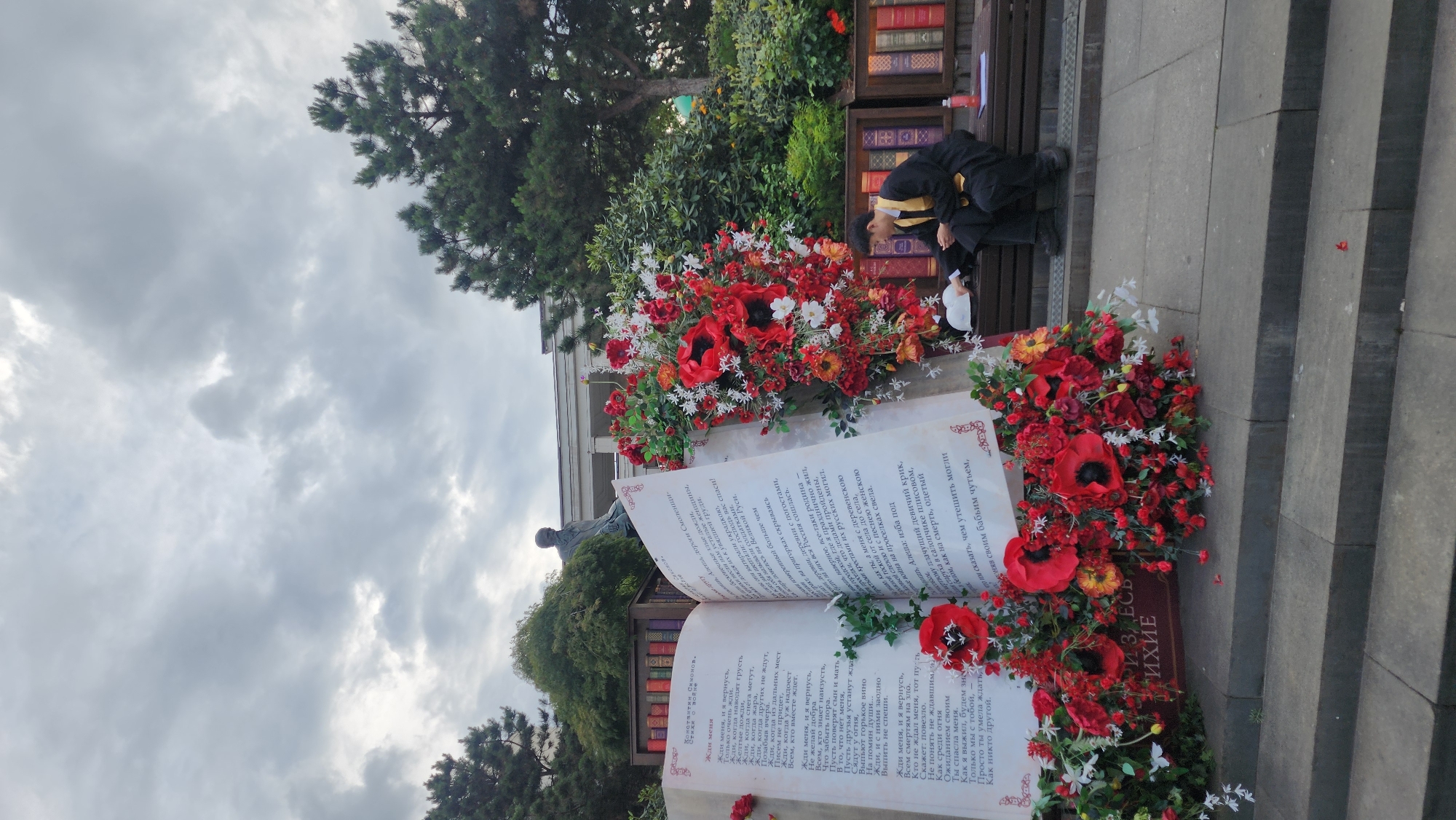The image depicts an outdoor scene in Moscow, Russia, specifically in the area of the Pushkin Literary Museum. In the foreground, a large open book is adorned with red poppies and white flowers. The book appears to be an art installation, with text visible on its pages. To the right, a young man dressed in a black and gold robe is seated on a bench, looking down at something in his hands. Behind the book and the seated man, a statue of Alexander Pushkin stands, overlooking the scene. The background features trees, some of which are evergreens, and a cloudy sky. Bookshelves filled with books are visible behind the statue and along the back of the bench area. The overall atmosphere suggests a memorial or cultural event related to literature.