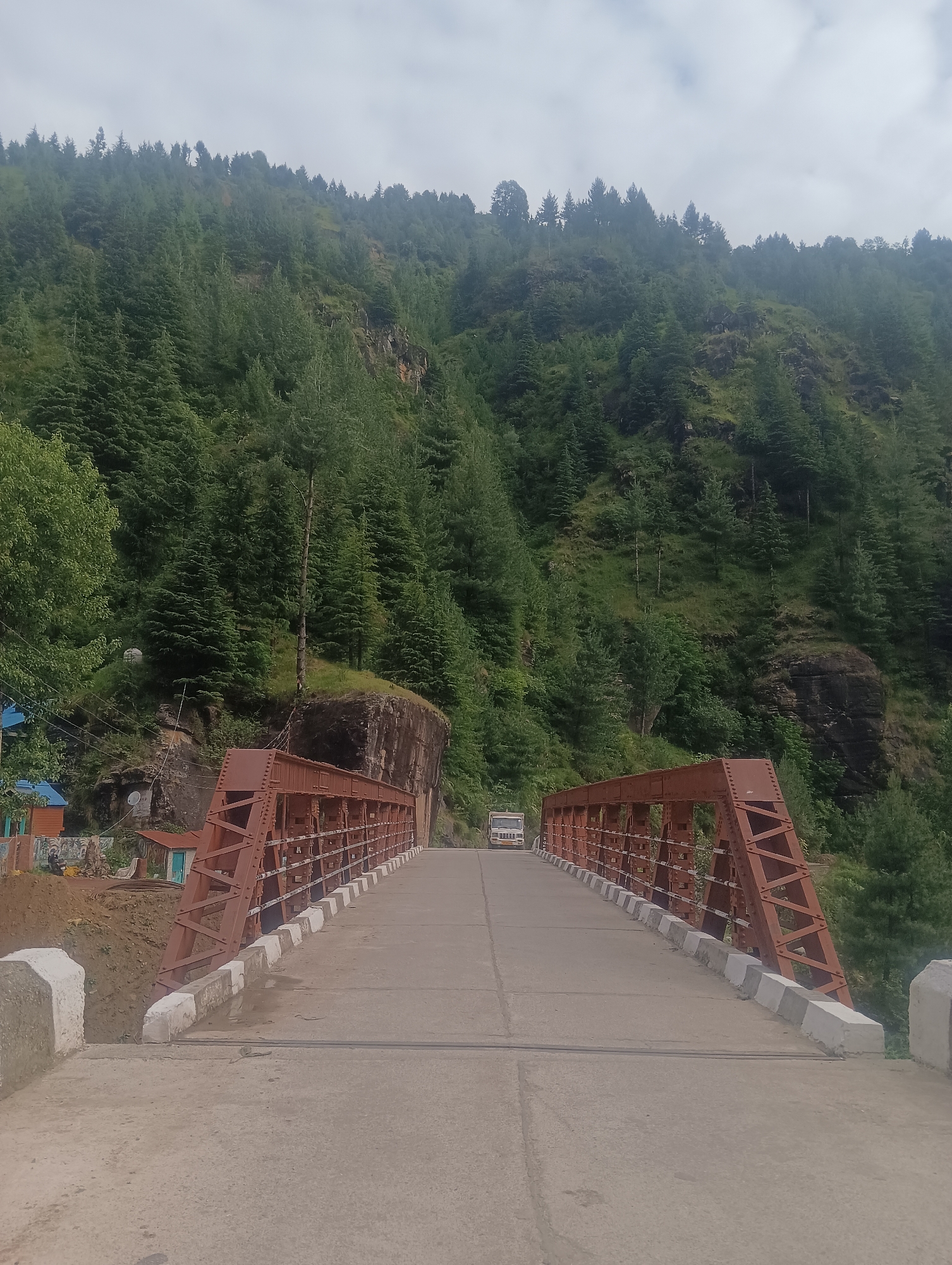 This is a full, eye-level, outdoor, vertical shot of a bridge in Shanag, India. The bridge is constructed of rusty red metal trusses and has a concrete road surface. On the bridge, a white truck is visible in the distance. The bridge spans a gap between two densely forested hillsides. The hills are covered in numerous pine trees and the sky is overcast. A small building with a blue roof is visible on the left side of the image, near the base of the bridge.
