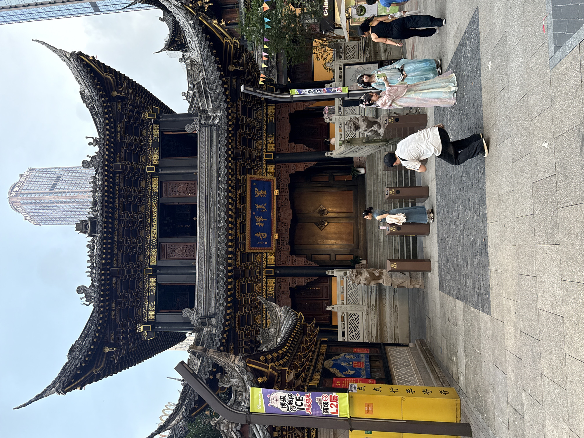 The image depicts a street scene in Yuzhong District, China, with a prominent traditional Chinese temple building as the central focus. The temple features intricate dark wooden architecture with ornate gold detailing and sweeping, curved roofs adorned with decorative figures. Two large wooden doors are visible at the entrance, set within a stone courtyard.
In the foreground, several people are present. A man in a white shirt and black pants is crouching down, seemingly taking a photograph with his phone. Two women are standing to his right, one wearing a pale pink and blue traditional Chinese dress, and the other in a light blue traditional Chinese dress. They are both looking at a phone held by the woman in the pink dress. Further to the right, a woman in a black tank top and black pants is walking away from the camera. On the steps of the temple, another woman in a blue dress is standing.
To the left of the temple, a tall yellow sign with Chinese text and colorful banners is displayed. A tall, modern skyscraper is visible in the background, juxtaposed with the traditional architecture. The ground is paved with large stone tiles and some darker cobblestones. Colorful triangular flags are strung across the scene above the temple entrance. The overall atmosphere suggests a busy urban environment with a blend of historical and modern elements.