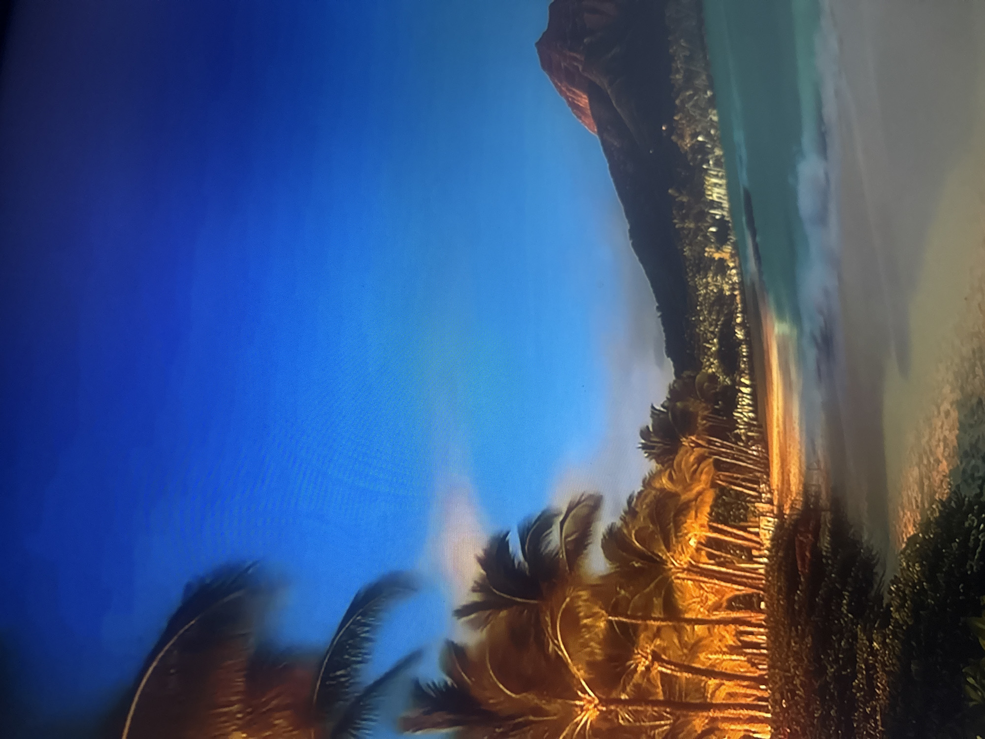 This is a photograph of a tropical beach scene, likely taken at sunset or sunrise. The foreground is dominated by the silhouettes of palm trees, their fronds illuminated by a warm, golden light. The trees are densely packed, creating a lush tropical feel. Beyond the trees, a strip of sand leads to the ocean, which appears calm with gentle waves lapping the shore. The water transitions from a light turquoise near the shore to a deeper blue further out.
In the background, a dark mountain or cliff face rises, its peak catching the last rays of the sun. The sky is a vibrant gradient of deep blue, transitioning to lighter hues as it approaches the horizon. There are some wispy clouds or mist present, adding to the atmospheric quality of the scene. The overall mood is serene and tranquil, evoking a sense of peace and natural beauty. There is no visible text in the image.