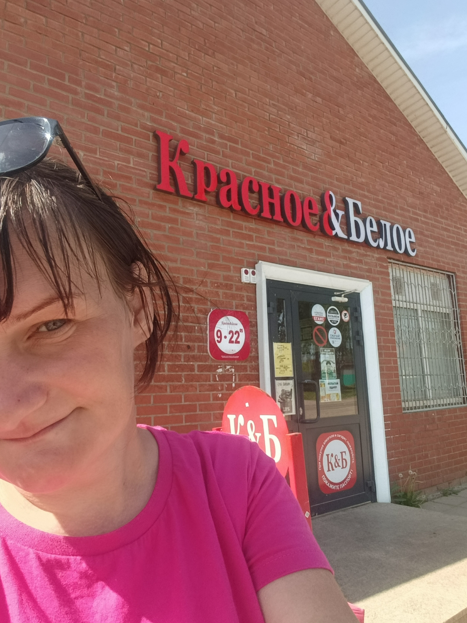 The image shows a woman wearing a pink shirt, posing in front of a brick building with a sign that says "Красное & Белое" in Russian. The sign is red and white. A sign on the front door says 9-22, referring to business hours. The image was taken in the city of Фоки, Russia.