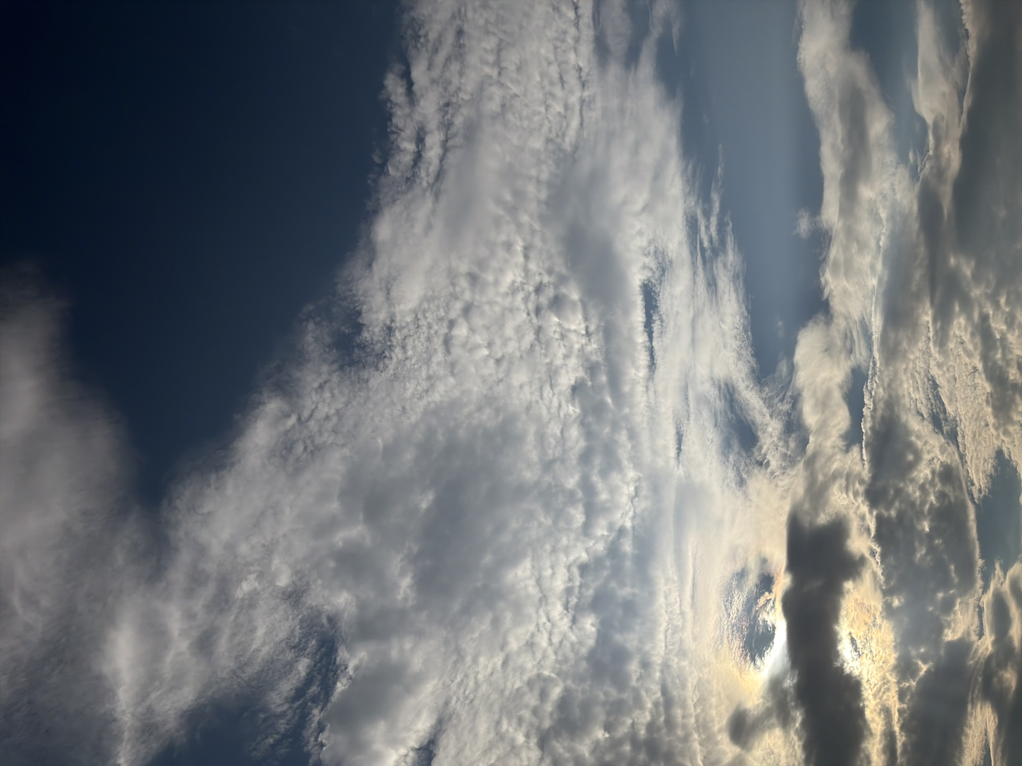 The image displays a sky filled with a variety of clouds, ranging from wispy cirrus formations to denser, more opaque cumulus clouds. Sunlight filters through the clouds in places, creating areas of bright illumination and casting shadows. The sky transitions from a deep, dark blue at the top to lighter shades of blue lower down. The scene appears to be an outdoor, daytime view of the sky, likely taken from ground level. There are no discernible elements of the city of Oxentea, Moldova visible in this sky-focused photograph.