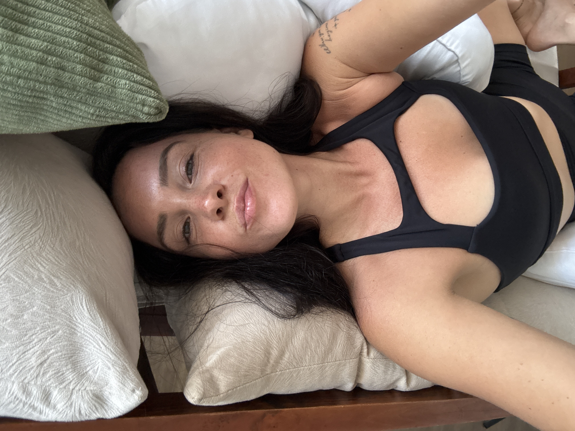 A woman with dark hair and freckles lies on her back, propped up by pillows. She is wearing a black sports bra and black shorts. A tattoo is visible on her upper arm. The environment appears to be indoors, possibly a bedroom, with a wooden frame visible on the left side of the image. The woman is looking directly at the camera with her lips pursed. The lighting is natural and soft.