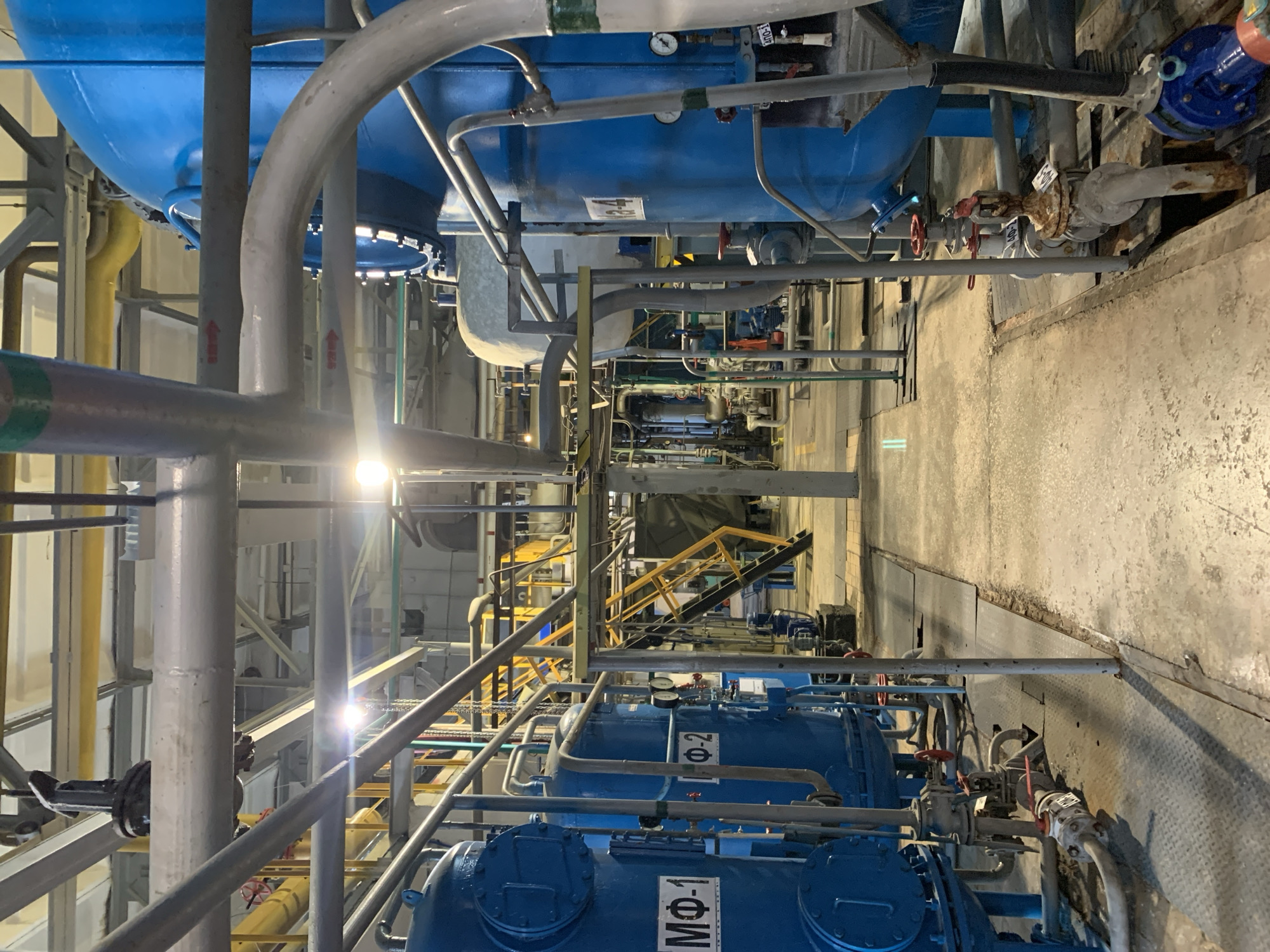 The image shows the interior of an industrial facility in what appears to be Chaikovsky, Russia, given the Cyrillic lettering on some of the blue machinery (МФ-1, Ф-2). The scene is dominated by an array of pipes, tanks, valves, and structural supports, suggesting a complex mechanical system. Most of the equipment is painted blue, with some pipes in yellow, gray, and silver. There is a catwalk with yellow handrails visible in the distance. The lighting is somewhat dim and industrial, with bright spots from light sources. There are no people immediately visible in the foreground of the image. The overall impression is of a functional, utilitarian space dedicated to some sort of industrial processing.