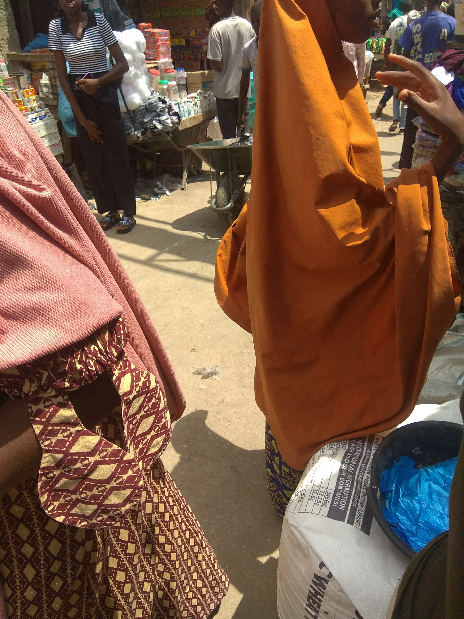 In a bustling outdoor market scene in Jalingo, Nigeria, several individuals are visible amidst stalls laden with goods. A young woman in a black and white striped shirt and black pants stands towards the left, holding a dark object. To her right, a person is partially obscured by a vibrant orange headscarf and garment, with an outstretched hand visible. In the background, other people can be seen, some wearing casual clothing, and a cart with various items. The ground is dusty, indicating a dry environment, and the lighting suggests it is daytime. A large white bag with visible text is positioned in the foreground, partially obscuring a black bowl filled with blue plastic bags. The text on the white bag reads "WHEAT IN PREMIA, ENZYME (LIPASE)" and provides nutritional information, including caloric content and macronutrient breakdown per kilogram. The overall impression is of a typical market day, with commerce and daily life unfolding in a lively atmosphere.