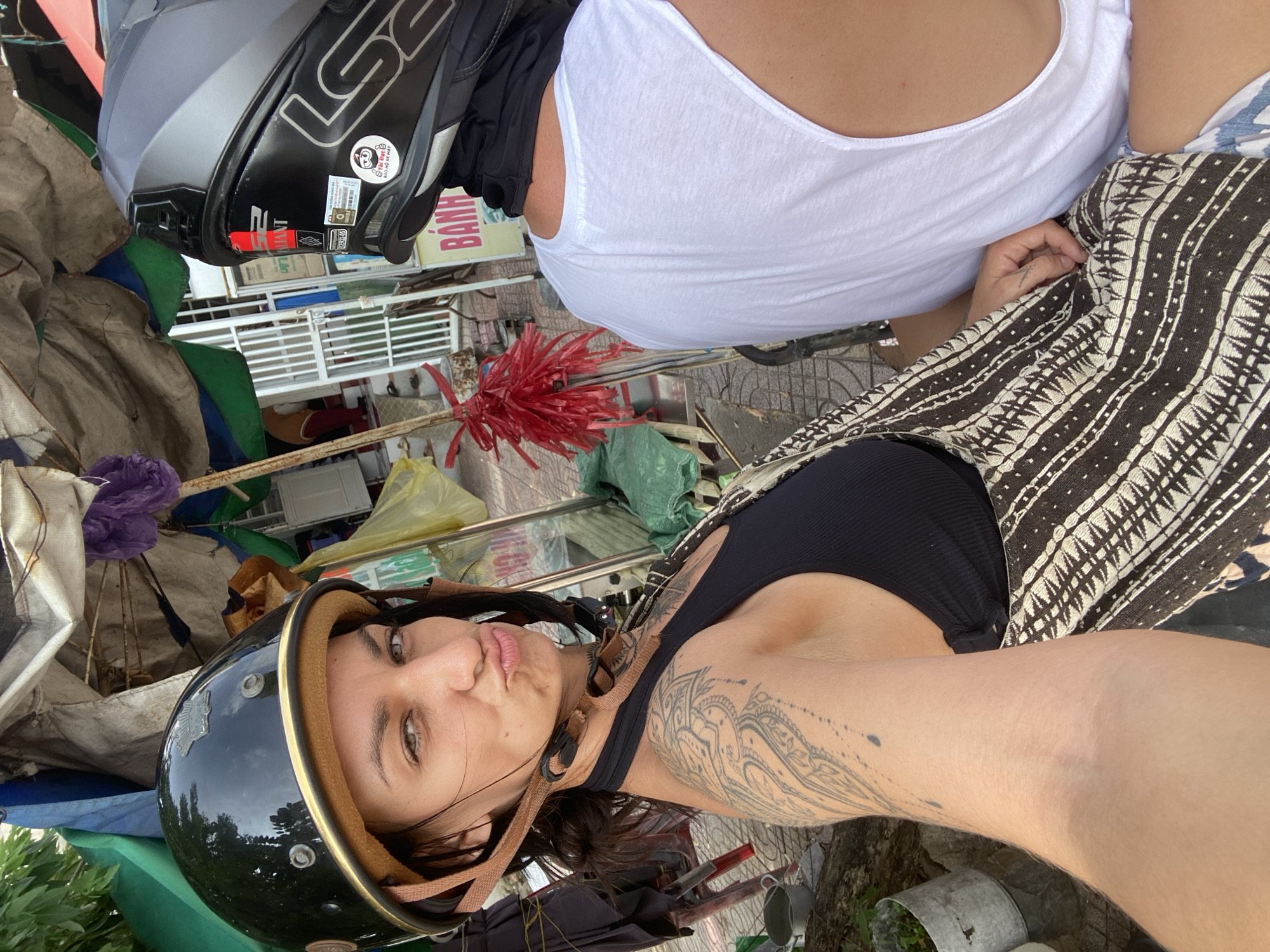 In Mũi Né, Vietnam, two individuals are captured in a selfie while seated side-by-side, likely on a scooter. The person in the foreground, a woman, wears a black helmet and a black tank top. She has a tattoo on her left arm. Her facial expression is a pout. The person behind her, wearing a white tank top and a black helmet, is partially visible. The environment appears to be a street scene with a makeshift shelter constructed from tarps and umbrellas in the background. Signs with Vietnamese text are visible, and a red plastic decoration hangs from a pole. The ground is paved, and some greenery and potted plants are present.
