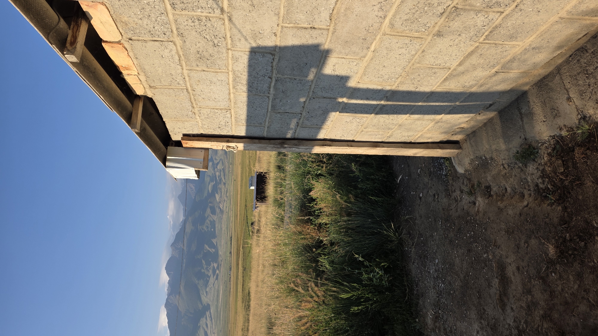 This vertical, medium shot photograph captures a rustic outdoor scene with mountains in the background, likely in the vicinity of Terek-Suu, Kyrgyzstan. In the foreground, the right side of the frame is dominated by a partially constructed wall made of light-colored concrete blocks. A wooden post is affixed to the wall, and a shadow cast by an unseen object stretches across the textured blocks.

To the left of the wall, a patch of dry, tall grass and other vegetation grows sparsely over a dirt ground. Further into the background, a flat, golden field stretches out, dotted with small, dark structures. In the distance, a range of majestic mountains, their peaks softened by the haze of the late afternoon or early morning sun, form the backdrop against a clear, bright blue sky. The overall impression is one of a rural, undeveloped area, possibly on the outskirts of a settlement.