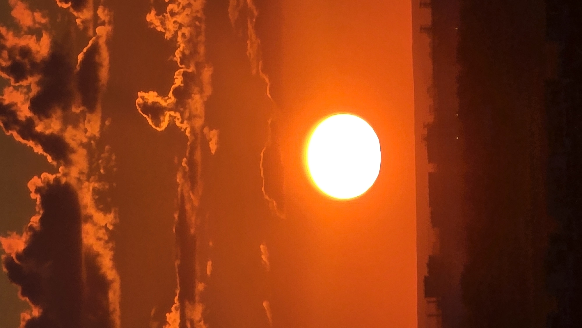 This is a vertical, eye-level shot of a sunset over Tolyatti, Russia. The sun, a bright white orb with a halo, is positioned slightly left of center in the middle of the frame. The sky is a vibrant orange, filled with fluffy, incandescent clouds that appear to be lit from behind by the sun. A silhouetted cityscape stretches across the bottom of the image, with buildings and trees faintly visible against the twilight. The overall impression is one of a serene and dramatic natural spectacle.