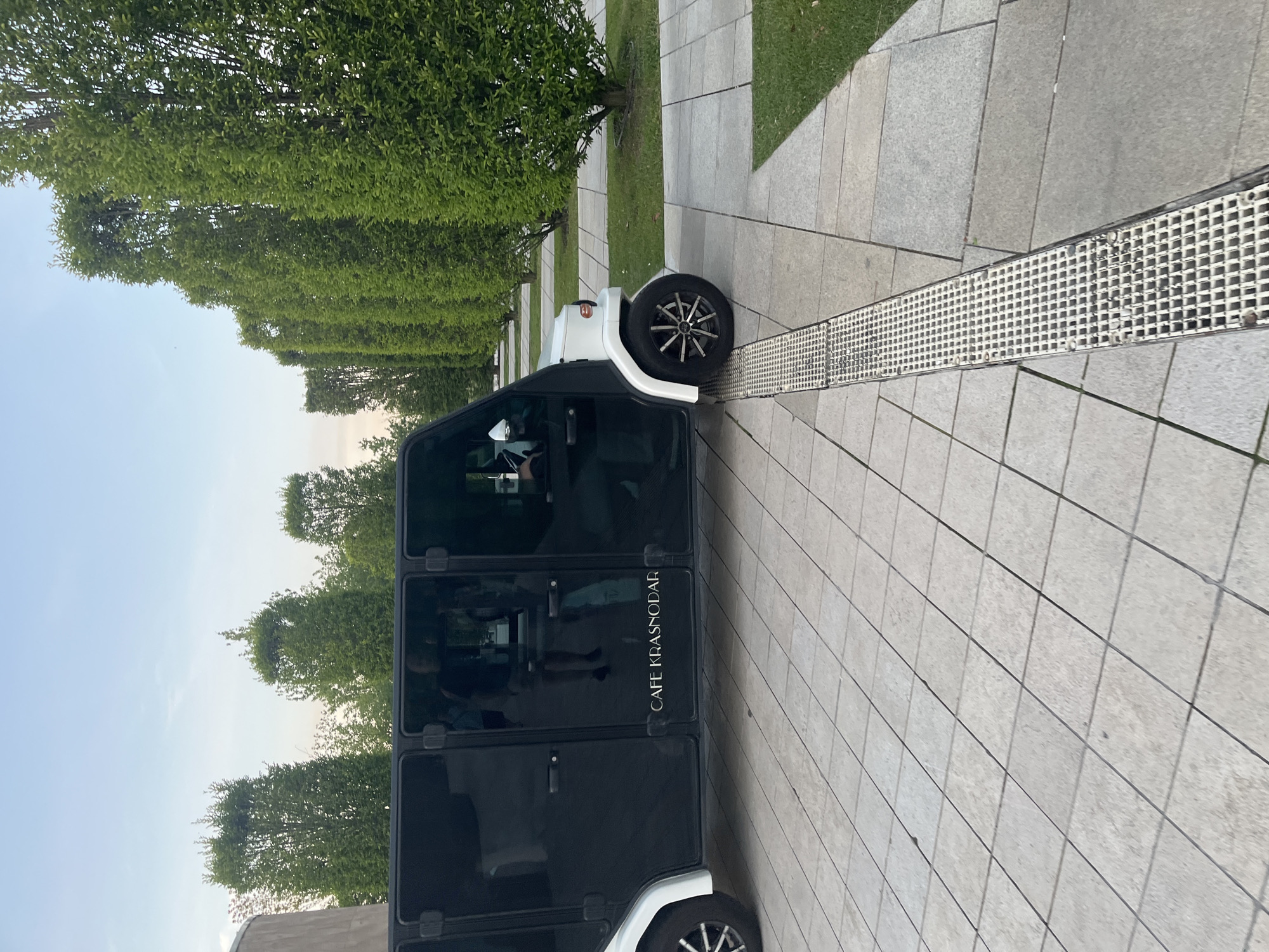 A small, white and black vehicle is parked on a tiled sidewalk next to a row of neatly trimmed green hedges. The vehicle appears to be a mobile cafe, with "CAFE KRASNODAR" visible in white lettering on its side. The tires of the vehicle are black with silver rims. A metal grate runs along the edge of the sidewalk, separating it from a grassy area. In the background, more hedges and trees are visible under a pale blue sky, suggesting it is daytime with clear weather. The scene is calm and orderly, with no specific activity or event clearly depicted, other than the presence of the vehicle itself. The location cues, such as the name on the vehicle and the implied setting of Krasnodar, Russia, are notable details.