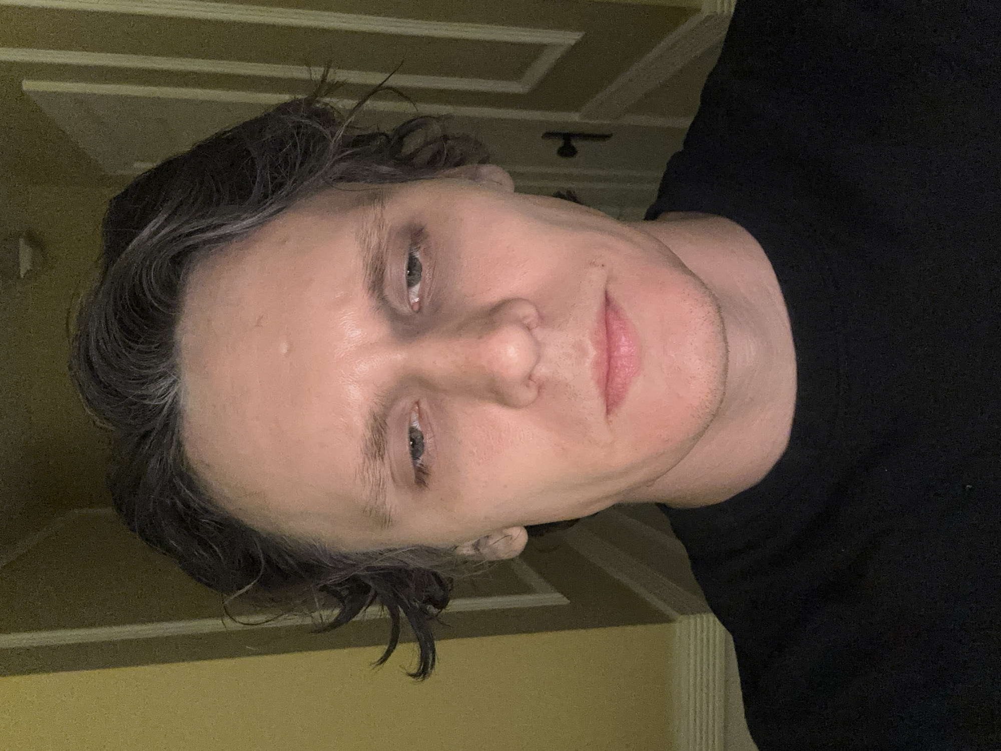 A medium shot portrait of a Caucasian male with dark hair and pale skin is shown. He is wearing a black t-shirt and is looking directly at the camera with a neutral expression. The background appears to be an indoor setting, possibly a hallway, with yellow walls and paneled doors. There is a faint shadow cast on the wall behind him, suggesting indoor lighting. The image is taken from a slightly low angle. The subject's facial features are prominent, with visible pores and some slight stubble on his chin. His hair is styled loosely, with some strands falling over his forehead. The lighting in the image is somewhat dim, creating a somber mood. There are no clear indications of the location within San Francisco, United States of America.