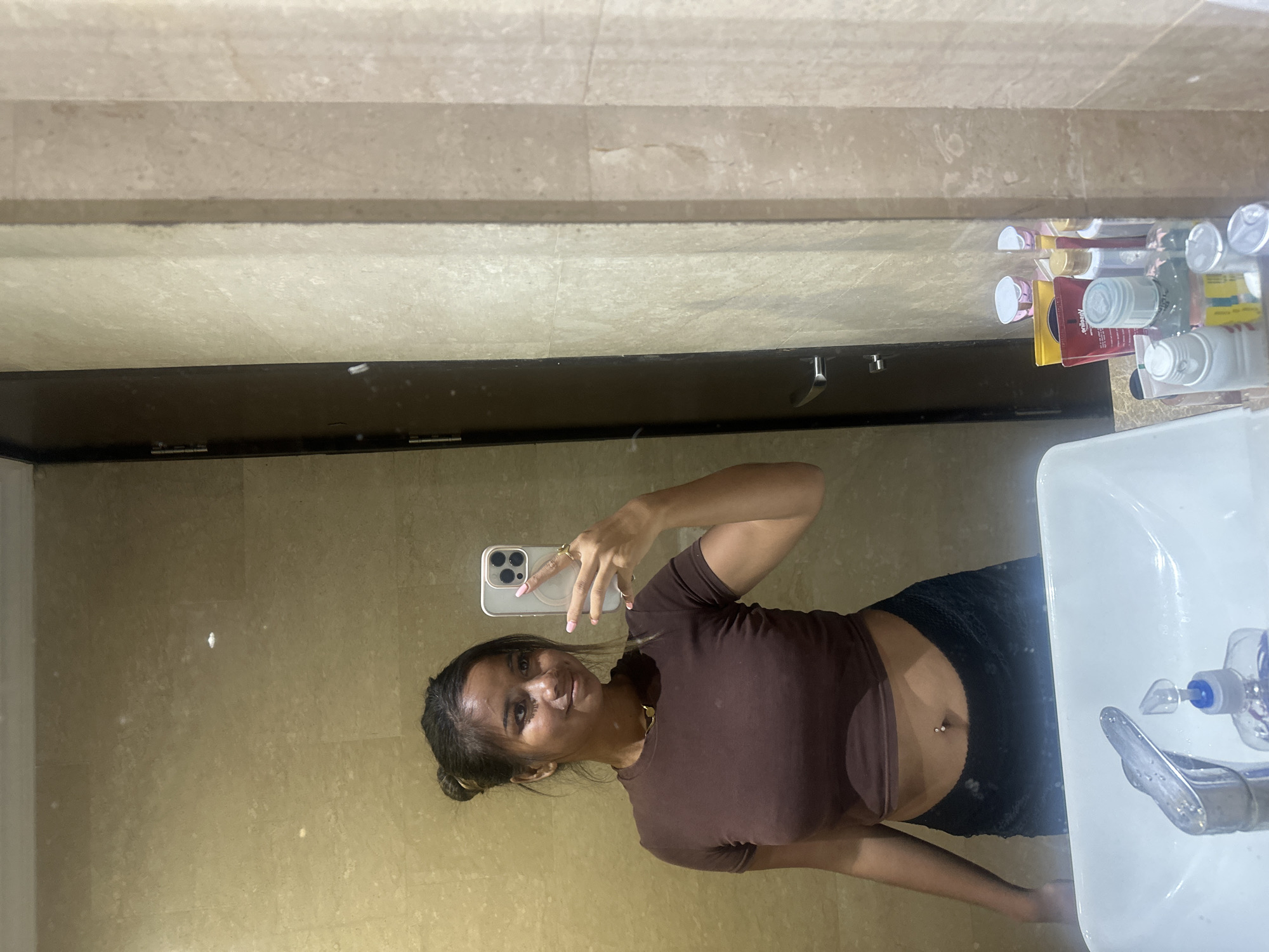 A woman is taking a selfie in a bathroom with beige tile walls and flooring. She is holding a silver iPhone with a triple-lens camera in her right hand. She has dark hair pulled back into a bun, and she is wearing a short-sleeved brown top and black shorts. A sink with a chrome faucet and soap dispenser is visible to the right, and various toiletries are displayed on a shelf above the mirror. The location is in Central Jakarta, Indonesia.