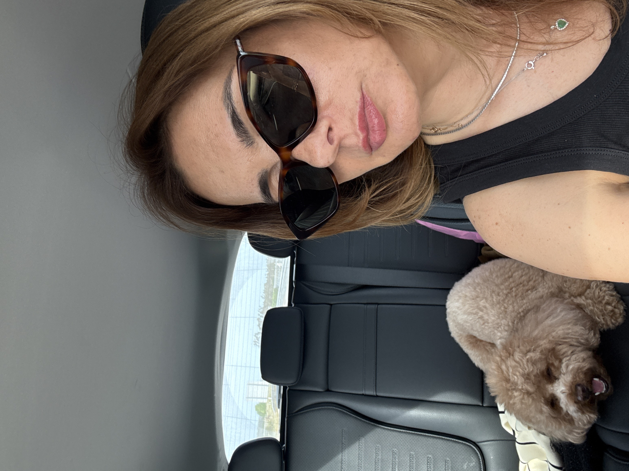 An upward-angled view captures a woman and a small dog inside a car, likely a selfie taken by the woman. The woman, positioned on the left, has shoulder-length, light brown hair. She wears large, tortoise-shell framed sunglasses that obscure her eyes, and her lips are softly parted with light pink lipstick. She is dressed in a black ribbed top and accessorized with two delicate silver necklaces, one featuring a small heart-shaped green pendant, and a ring on her finger.
To her right, a fluffy, light brown dog, resembling a poodle, rests on the black leather car seat. Its mouth is slightly open, suggesting it might be sleeping or yawning. A striped piece of fabric is visible beneath the dog.
The car's interior features black leather seats and headrests. Through the car window in the background, a bright daylight scene is visible, hinting at clear weather. The distant landscape appears open, possibly rural or suburban, but specific details are indistinct. The overall scene suggests a relaxed moment, possibly during travel in Pombal, Portugal. No visible text is present in the image.