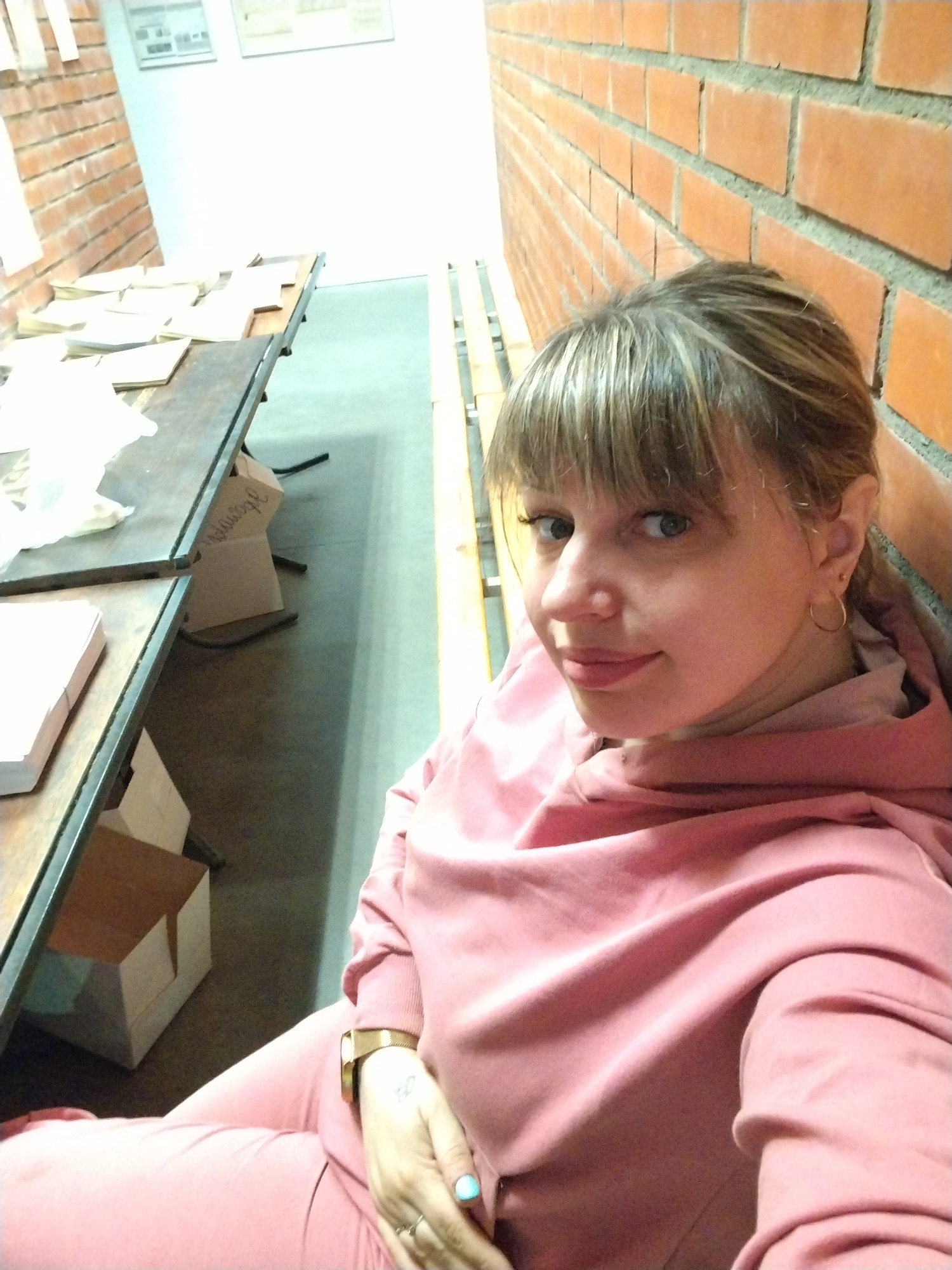 The image features a young woman taking a selfie indoors. She has light-colored hair with bangs, light eyes, and a neutral, slightly smiling expression. She is wearing a pink hooded tracksuit. Notable accessories include a watch on her left wrist, small hoop earrings, and a light blue painted fingernail on her right hand. A tattoo is also visible on her left forearm.
The woman is positioned near a reddish-orange brick wall on the right side of the frame. To her left, a long, dark-colored table or workbench is visible, laden with stacks of papers or books. Several cardboard boxes are placed underneath this table. The background on the left reveals another brick wall and a white wall displaying framed documents or pictures. The floor appears to be concrete or industrial flooring, and a yellow or light-colored railing runs along the floor on the right.
The setting suggests an indoor workspace, such as an office, workshop, or perhaps an archive or library, characterized by its functional layout and brick architecture. No specific activity other than posing for the photograph is evident. The word "Sudoge" (Сyдоге) or "Sudogel" is partially visible in Cyrillillic script on one of the cardboard boxes under the table. The time of day is not discernible, as the scene is indoors, likely under artificial lighting. The woman appears calm and relaxed. The context provided specifies the location as Rusanovo, Russia.