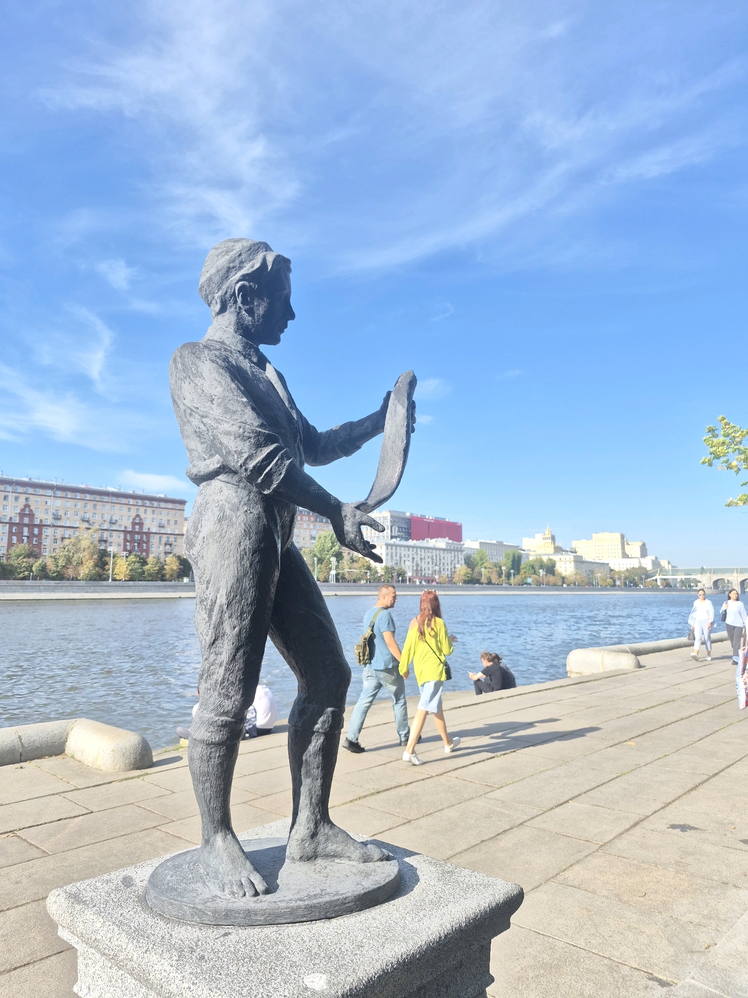 The image depicts a bronze statue of a man holding a scroll on a granite pedestal in Moscow, Russia. The statue is located on a paved embankment beside a wide river, likely the Moskva River, with buildings visible across the water. Several people are walking along the embankment, enjoying the sunny day with a clear blue sky and scattered clouds. The architecture of the buildings suggests a historical urban setting.