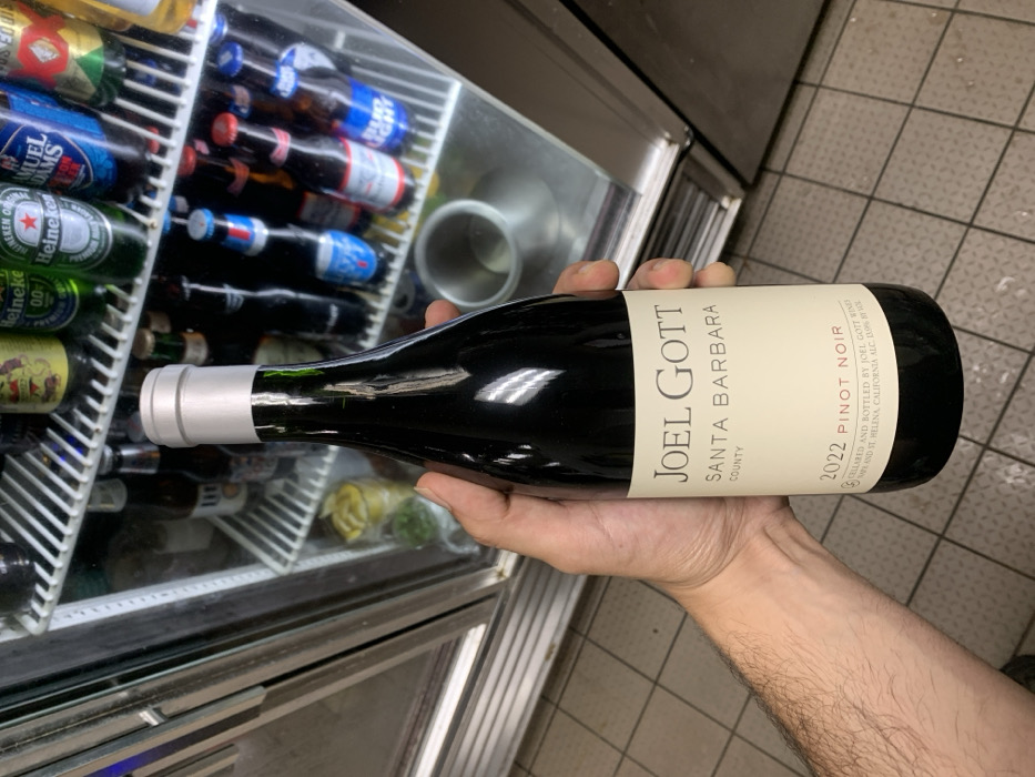 A person holds a bottle of "Joel Gott" Santa Barbara County Pinot Noir from 2022. The bottle has a silver cap and a beige label. In the background, a refrigerated display case is stocked with beer bottles, including Heineken, Samuel Adams, and Bud Light. The background also reveals a tiled floor and part of a stainless steel counter. The wine bottle is the primary focus, held securely in the person's hand.