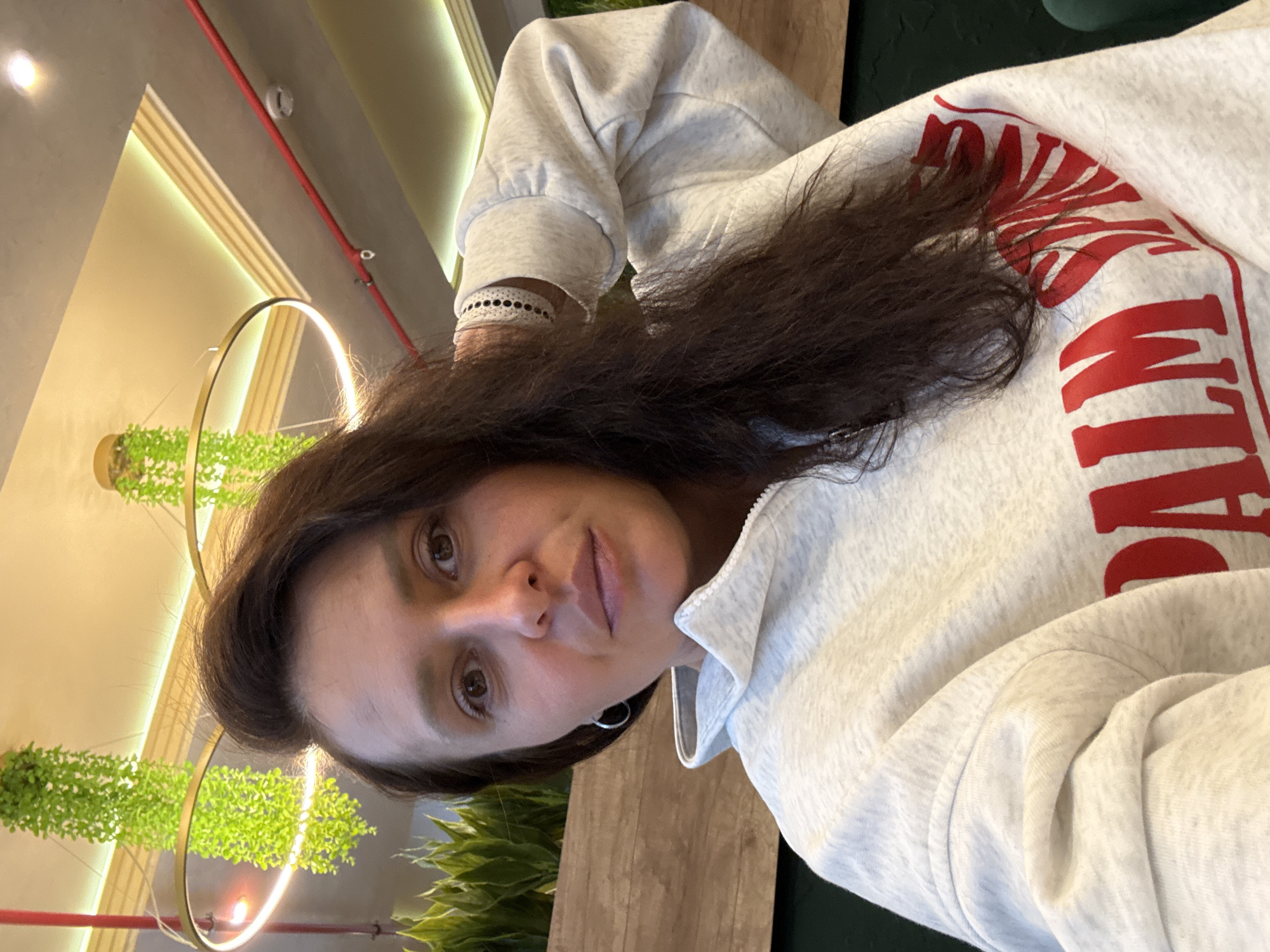 A woman with shoulder-length, dark brown hair is seated indoors. She is wearing a light grey hoodie with red lettering. The location appears to be a restaurant or cafe, as indicated by the decorative hanging plants with illuminated rings and wooden table. The image does not contain enough information to determine if the location is in Neftekamsk, Russia.
