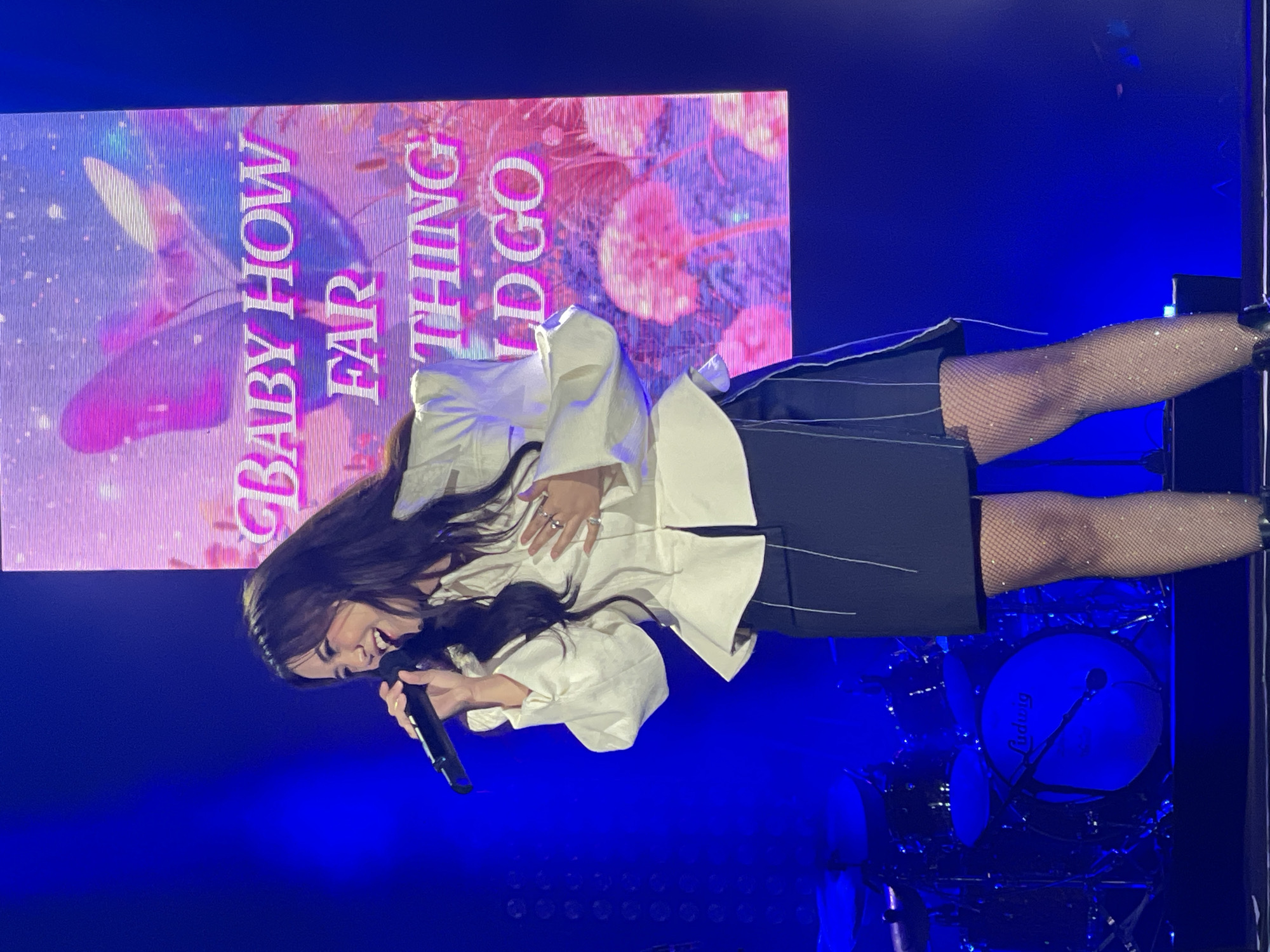 A woman is singing into a microphone on a stage with a drum set and an LED screen behind her that says "Baby How Far Things Go". The stage is lit with blue lights. The location appears to be a concert venue in Salembaran, Indonesia.